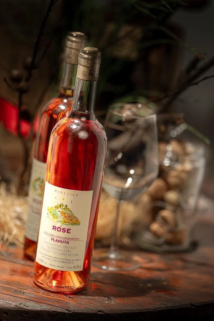 Two bottles of rosé wine on a wooden surface with a wine glass and decorative jar filled with corks in the background