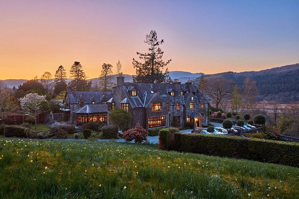 Penmaenuchaf Hotel is shown at sunset, surrounded by lush greenery, trees, and a mountain landscape in the background.