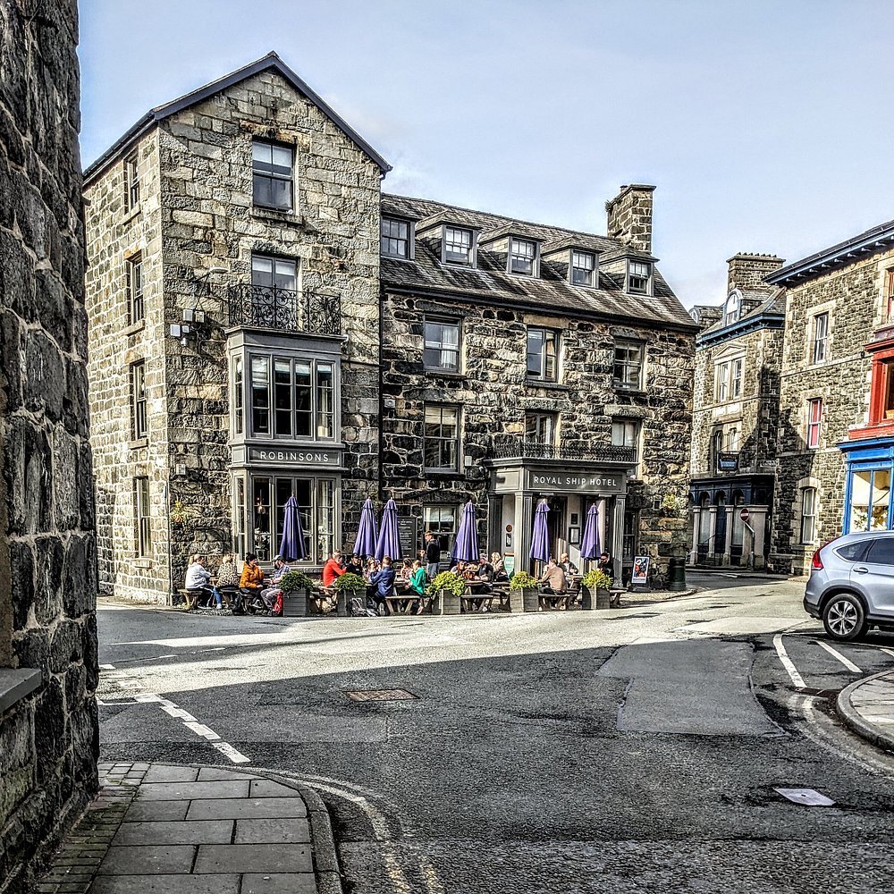 A stone building in Dolgellau called 'Royal Ship Hotel' with outdoor seating and purple umbrellas, people sitting at wooden tables, parked cars on the street, and other stone buildings in the background.