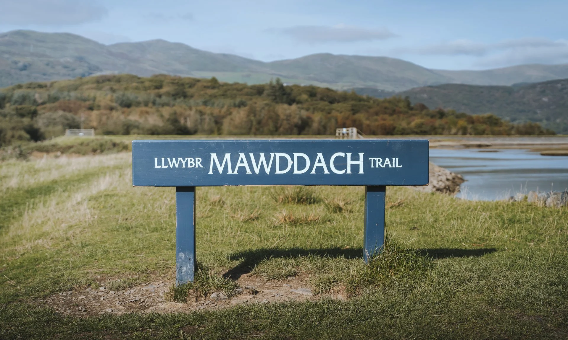 A sign that reads 'LLWYBR MAWDDACH TRAIL' in front of a landscape with hills, grass, and water, under a partly cloudy sky.