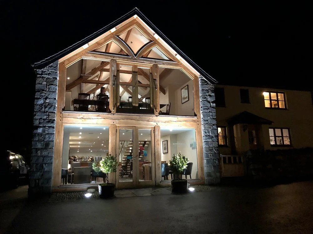Mawddach Pizza, illuminated at night. The upper floor is open with visible wooden beams and furniture, and the lower floor shows a well-lit interior with plants and seating.