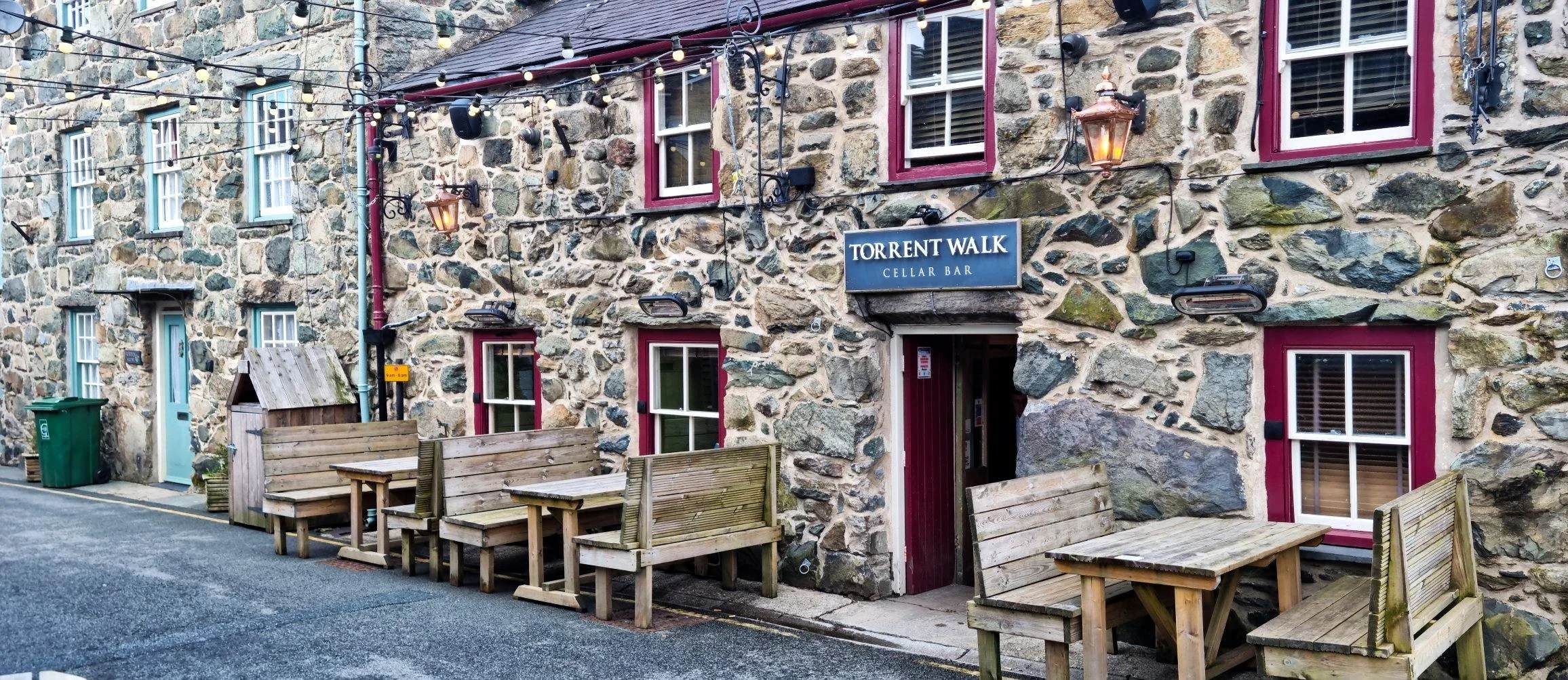 Exterior of a Dolgellau stone building with a blue sign that reads 'Torrent Walk Cellar Bar', red-framed windows, and outdoor seating with wooden benches and tables on a sidewalk, decorated with string lights and lanterns.