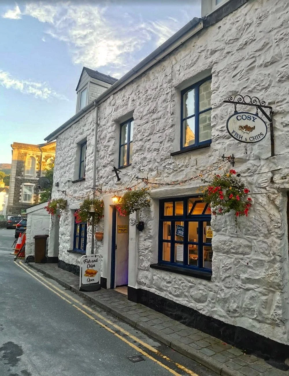 Cosy Fish and Chip shop in Dolgellau, it's a white stone building with blue trim, has hanging flower baskets.