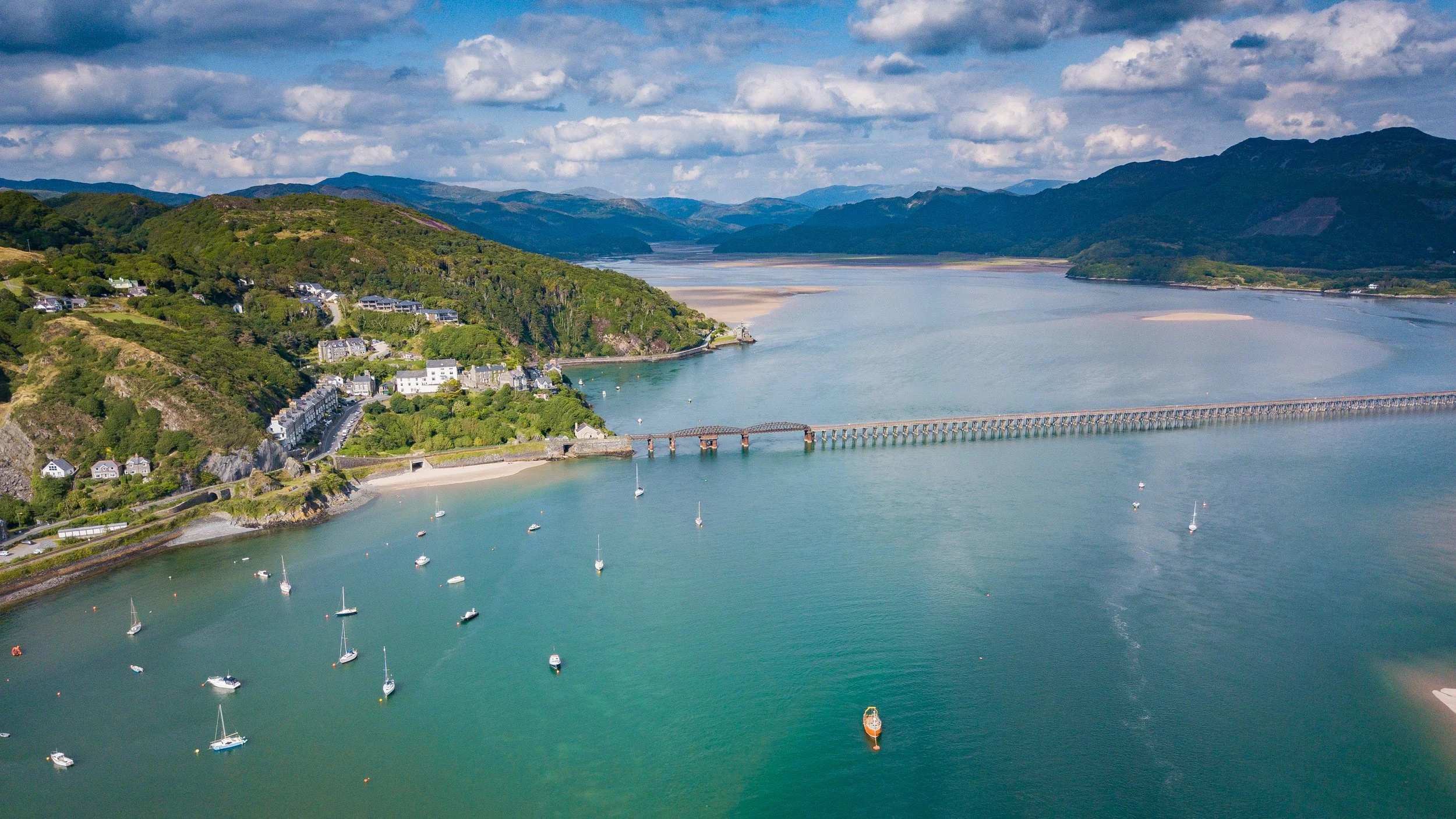 An aerial view of Barmouth, near Dolgellau with a bridge over water, boats anchored, green hills, and mountains in the background under a partly cloudy sky.