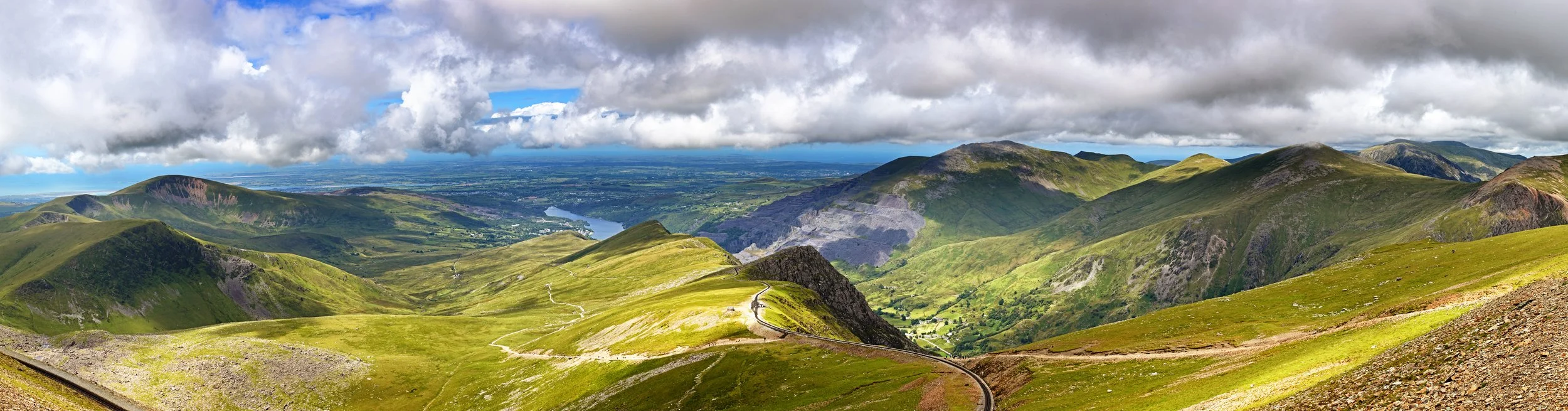Panoramic view of lush green mountains, winding roads, and a distant lake under cloud-filled sky.