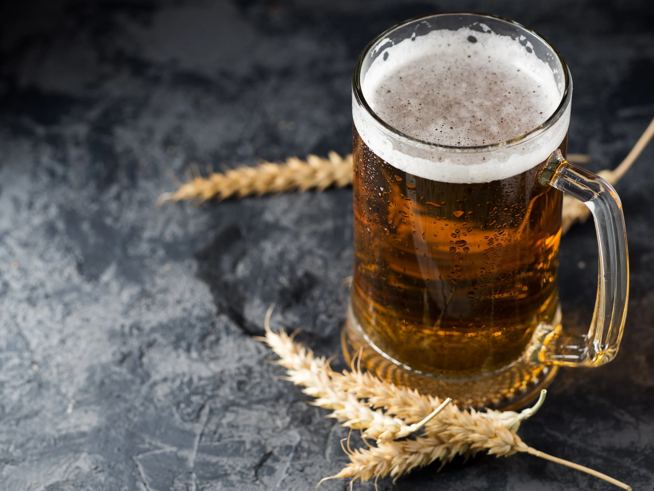 A glass mug filled with beer, showing condensation on the outside, placed on a dark textured surface with a wheat stalk beneath it.