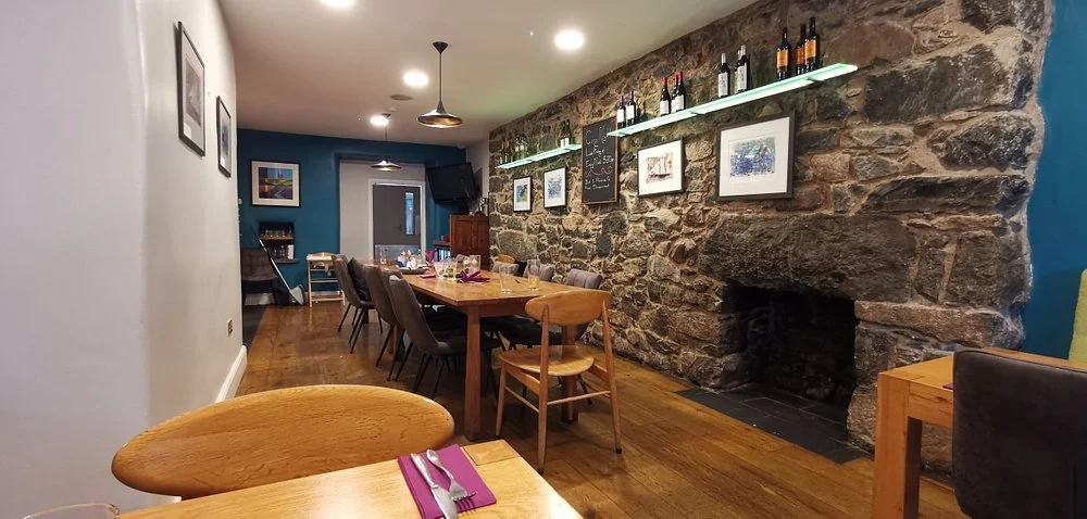 A cozy restaurant interior at the Cross Foxes Pub outside Dolgellau in the Snowdonia National Park, with a stone fireplace on a brick wall, framed art, a bar shelf with wine bottles, and a long dining table with chairs, set with utensils and napkins.