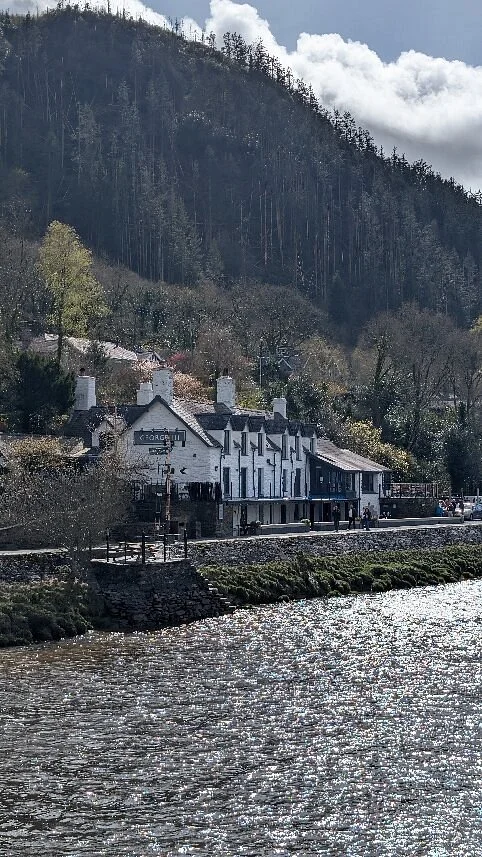 The George III in Snowdonia shows mountain with dense forest, a the pub with white walls and chimneys, and a walkway along the river.