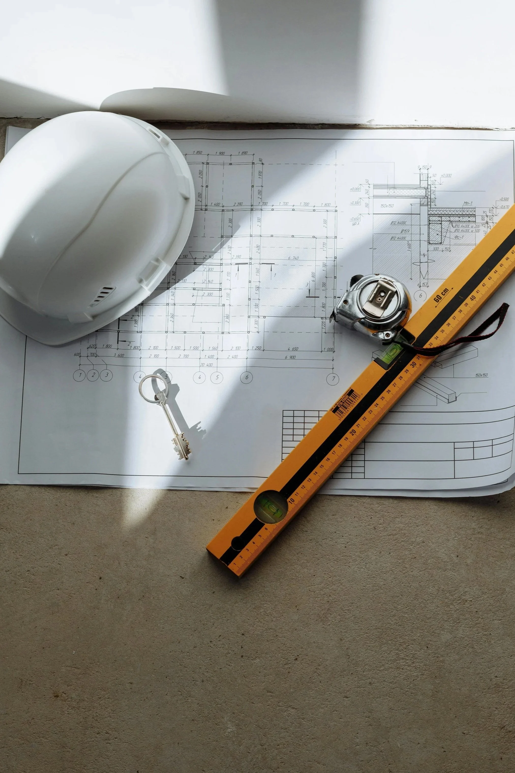 Blueprint drawings on paper with a white construction helmet, a metal measuring tape, a level, a yellow ruler, and a silver key, all laid out on a concrete surface.