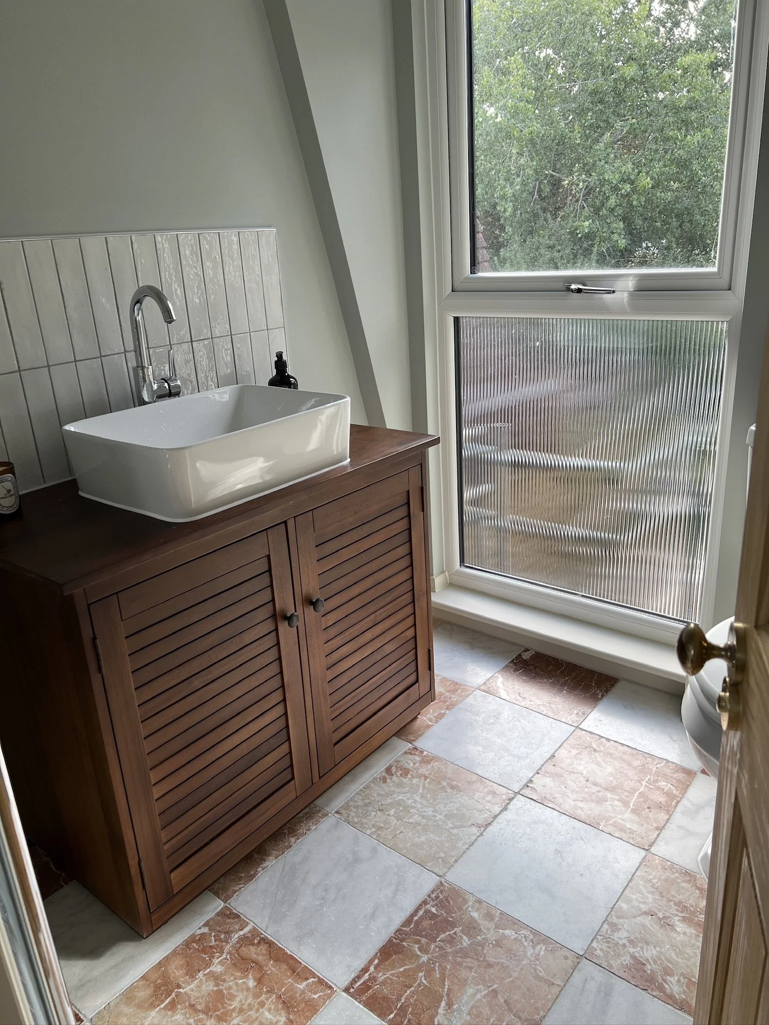 Bathroom with a wooden vanity with a white rectangular sink, a chrome faucet, a soap dispenser, a window with a view of green trees, and a tiled floor with beige and reddish tiles.