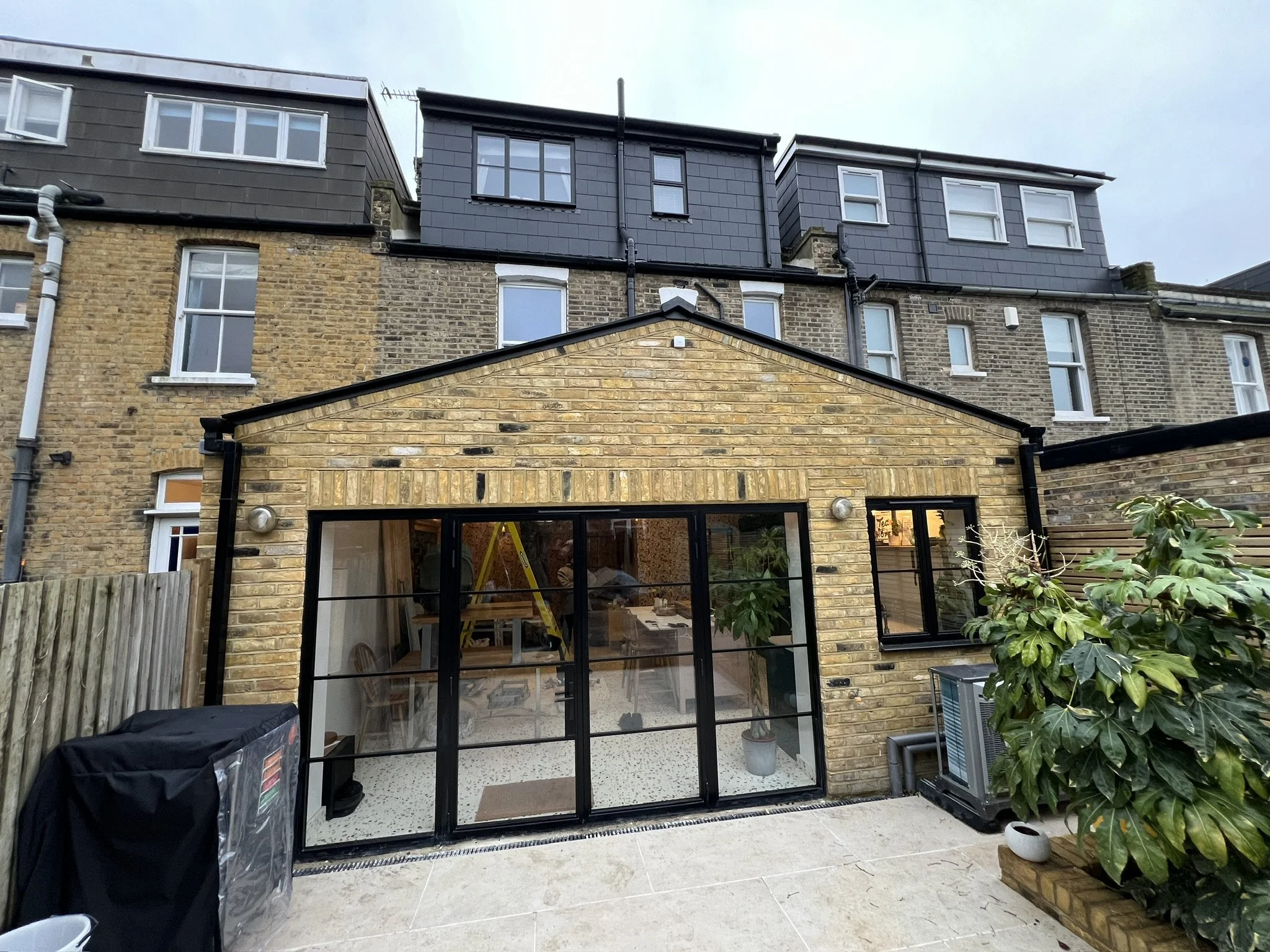 Backyard patio with a brick house, black metal-framed glass doors, potted plants, a gas grill covered with a tarp, and an air conditioning unit. The house has a brick lower level and a gray upper level with multiple windows.