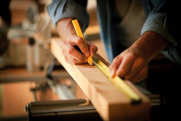 Person measuring a piece of wood with a yellow tape measure in a woodworking shop.