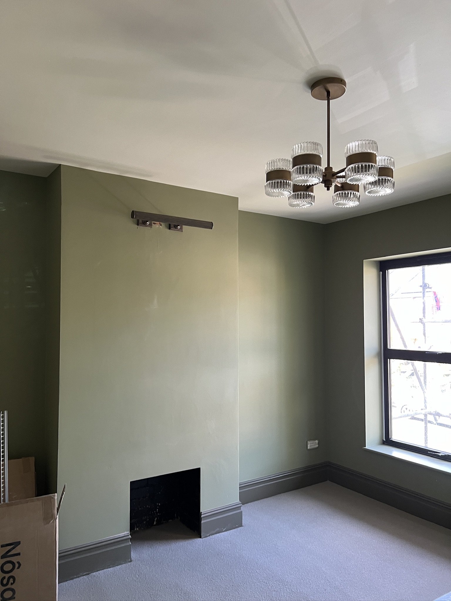 A room with green walls, a window, a chandelier, and carpet flooring. There is a small black fireplace vent and some boxes near the wall.