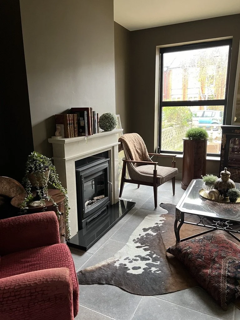 Cozy living room corner with a fireplace, a wooden armchair with a throw blanket, a red armchair, a glass-top coffee table with decorative items, and potted plants near a large window letting in natural light.
