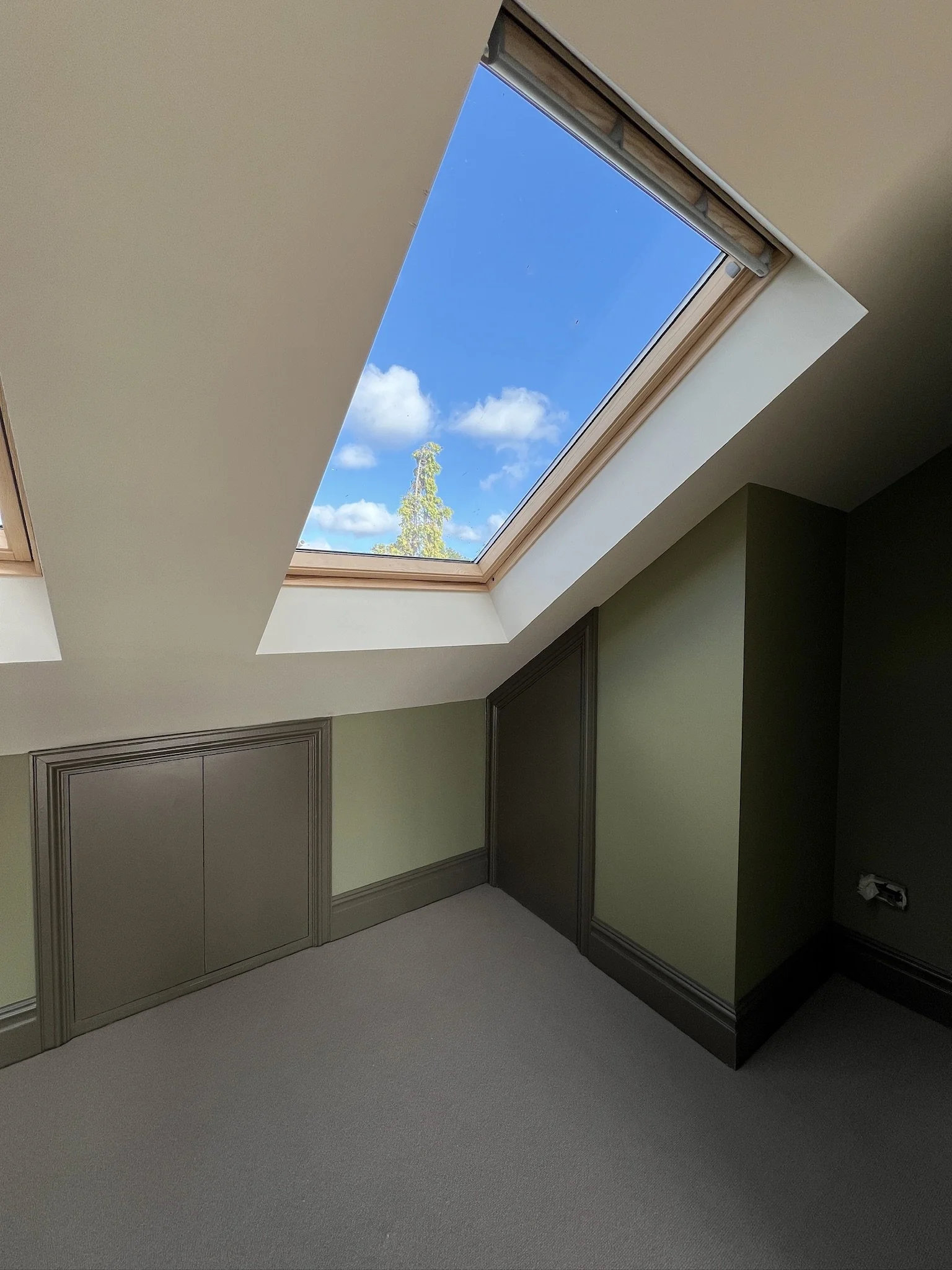 An attic room with a skylight window showing blue sky and some clouds, beige carpeted floor, green and dark gray walls, beige ceiling, and built-in cabinets.