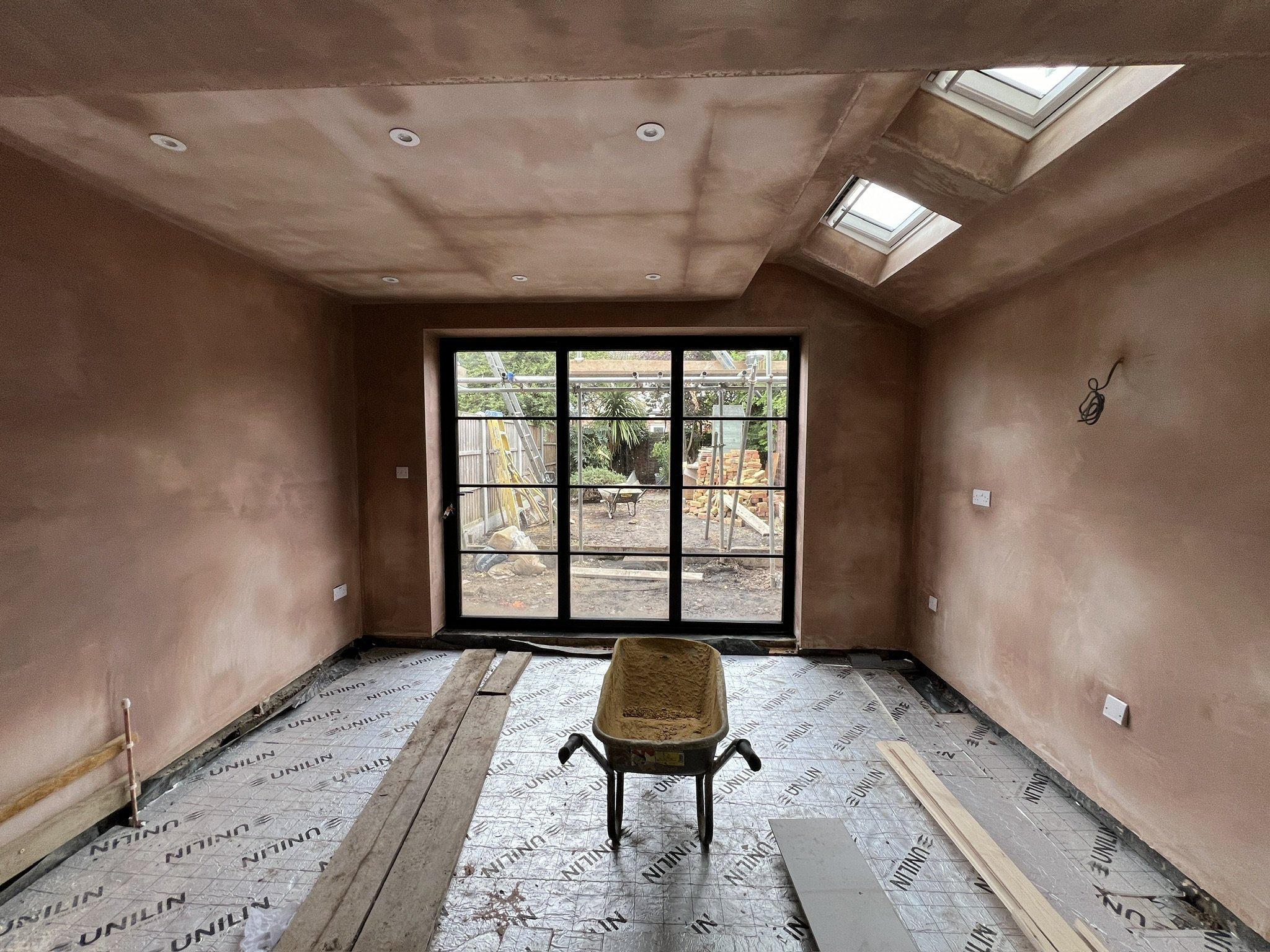Interior of a room under construction with a large window, skylights, and partially finished walls and ceiling, covered with protective sheets and construction materials.