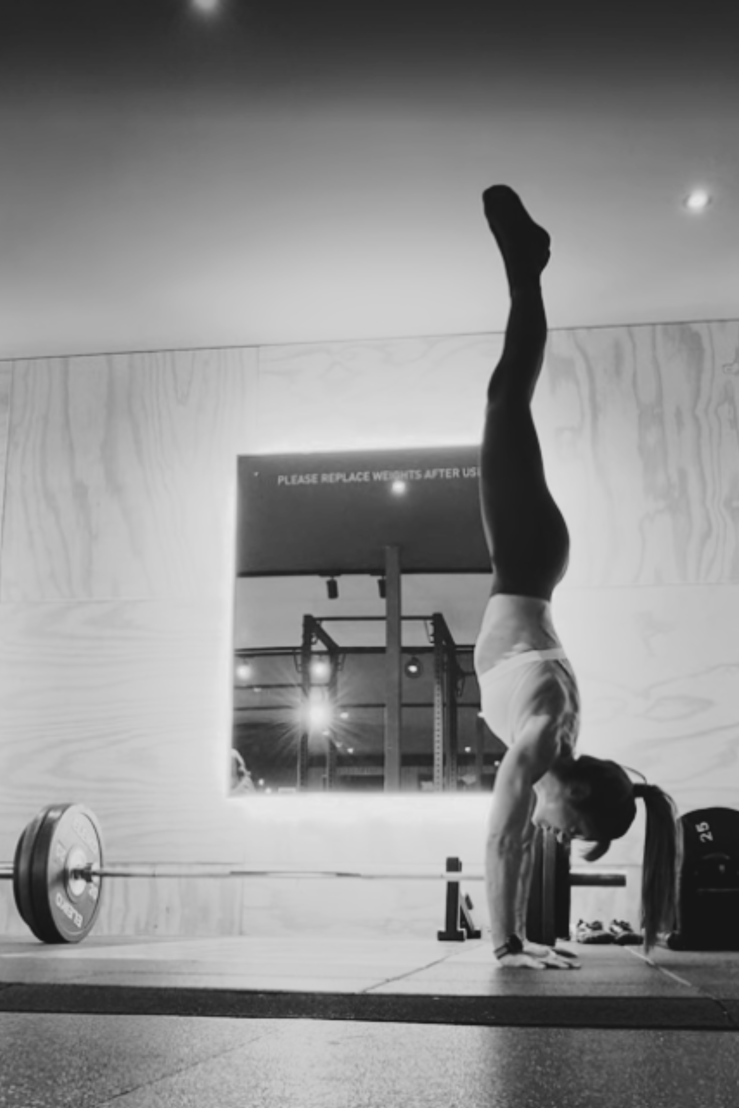 A woman doing handstand in a gym, with weights and gym equipment in the background, and a sign that says 'Please replace weights after use.'