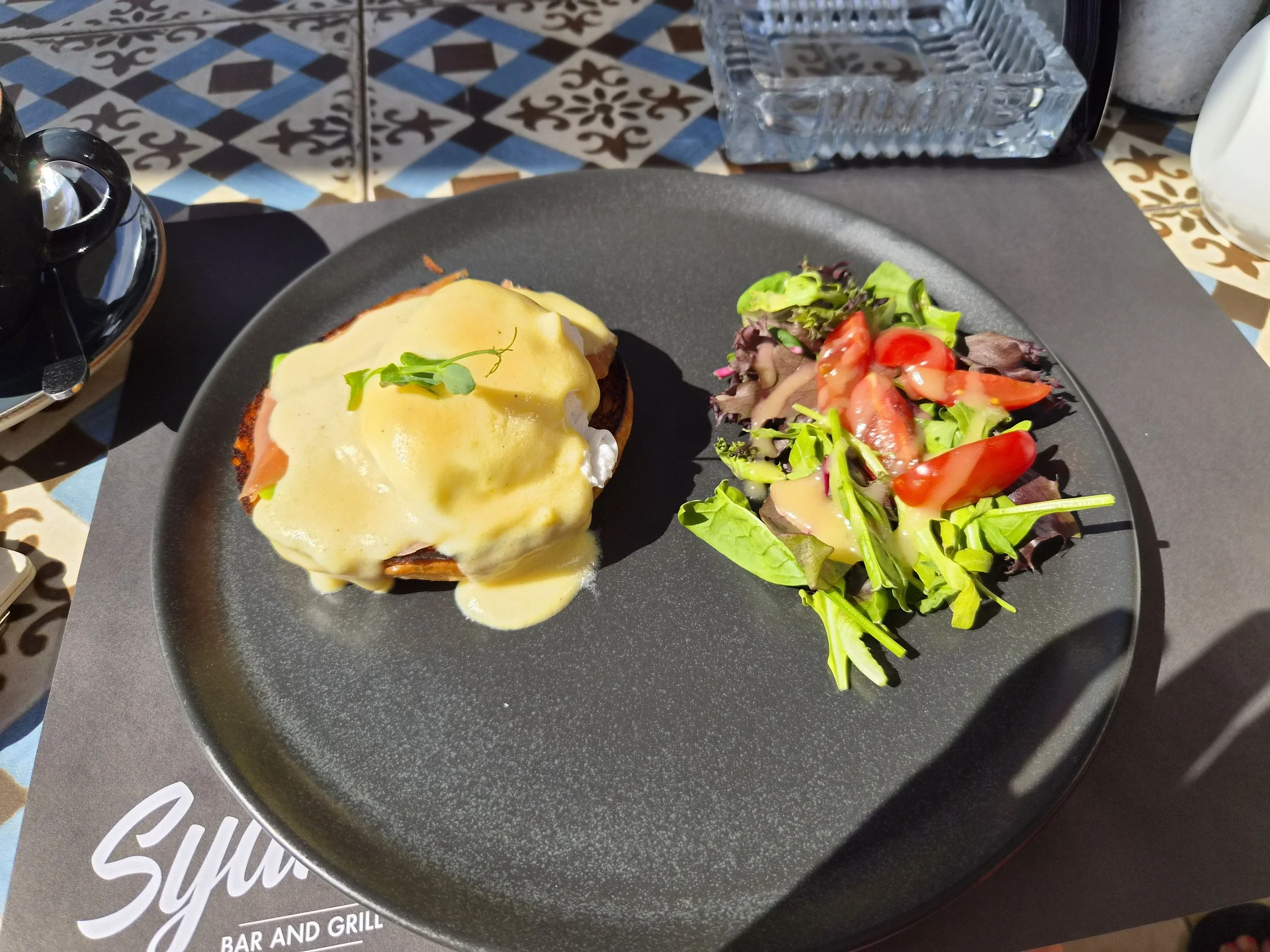 A black plate with eggs Benedict topped with hollandaise sauce, served with a side salad of mixed greens and cherry tomatoes on a table with colorful patterned tiles.