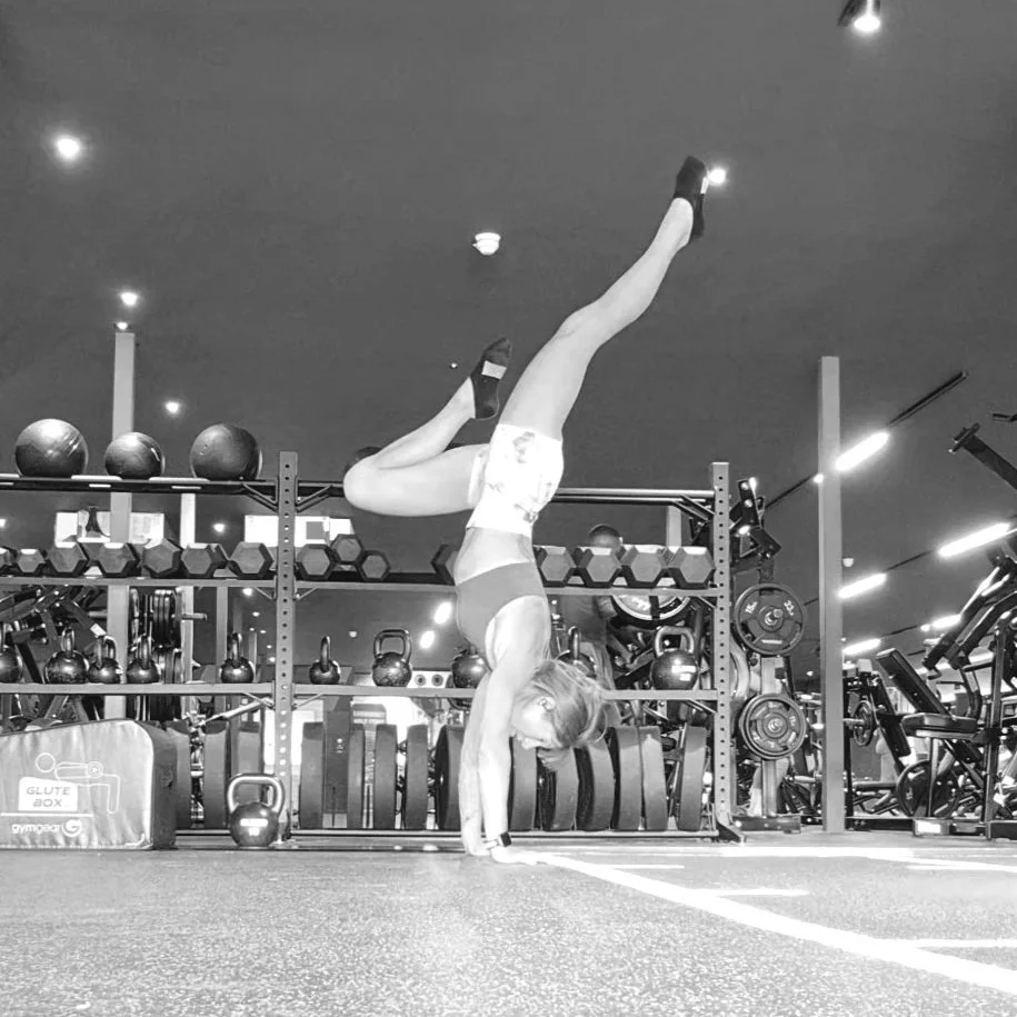 A woman performing a handstand in a gym, with one leg extended upward and the other bent. She is wearing sports bra, shorts, and sneakers, on a rubber gym floor with workout equipment in the background.