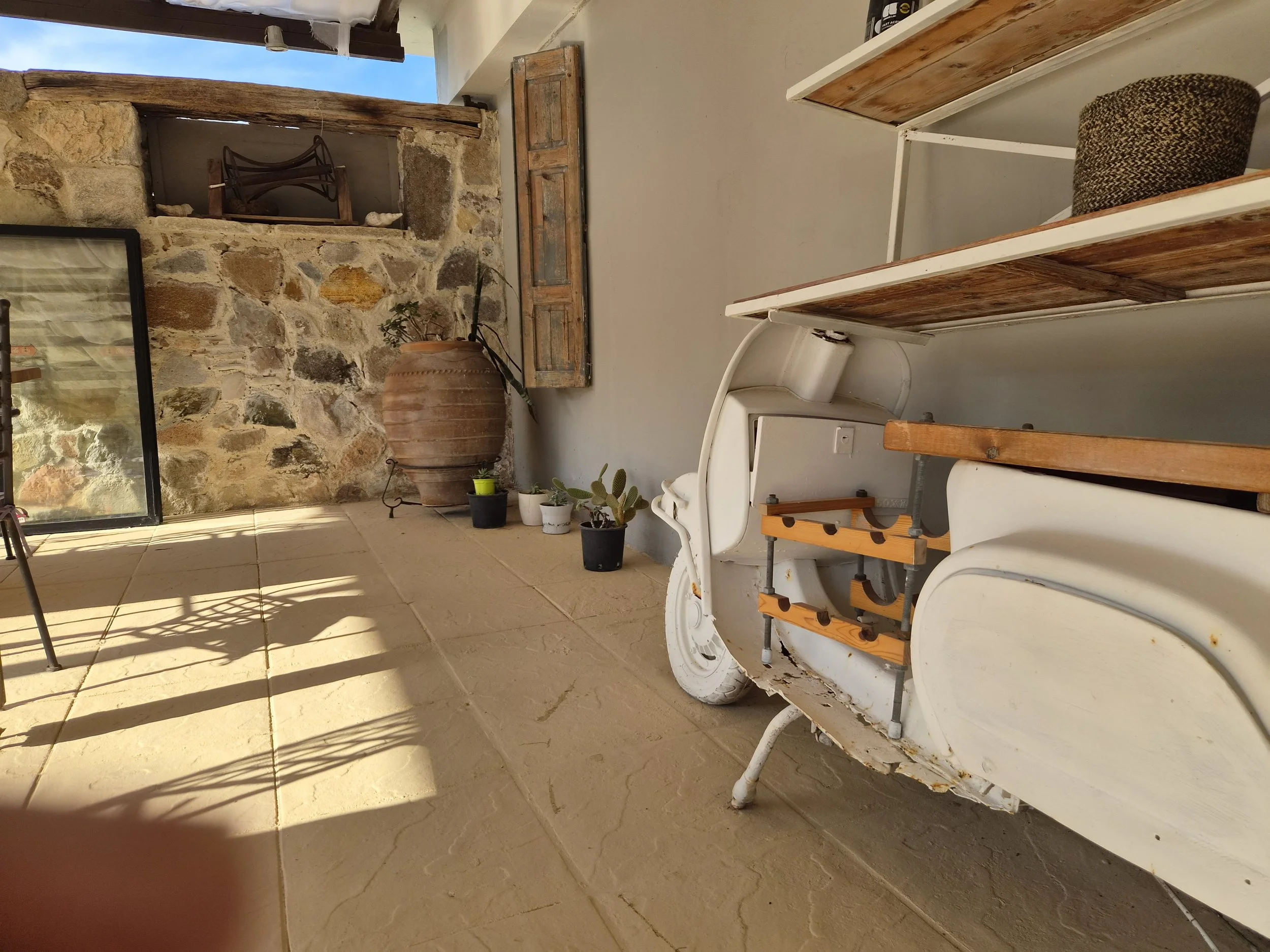 A rustic outdoor patio area with a stone wall, beige tiled flooring, and a white metal bench with wooden slats. There are several potted plants near the wall, a large clay pot, and decorative items on the stone ledge. Shadows from the railing are cast on the floor, indicating sunny weather.