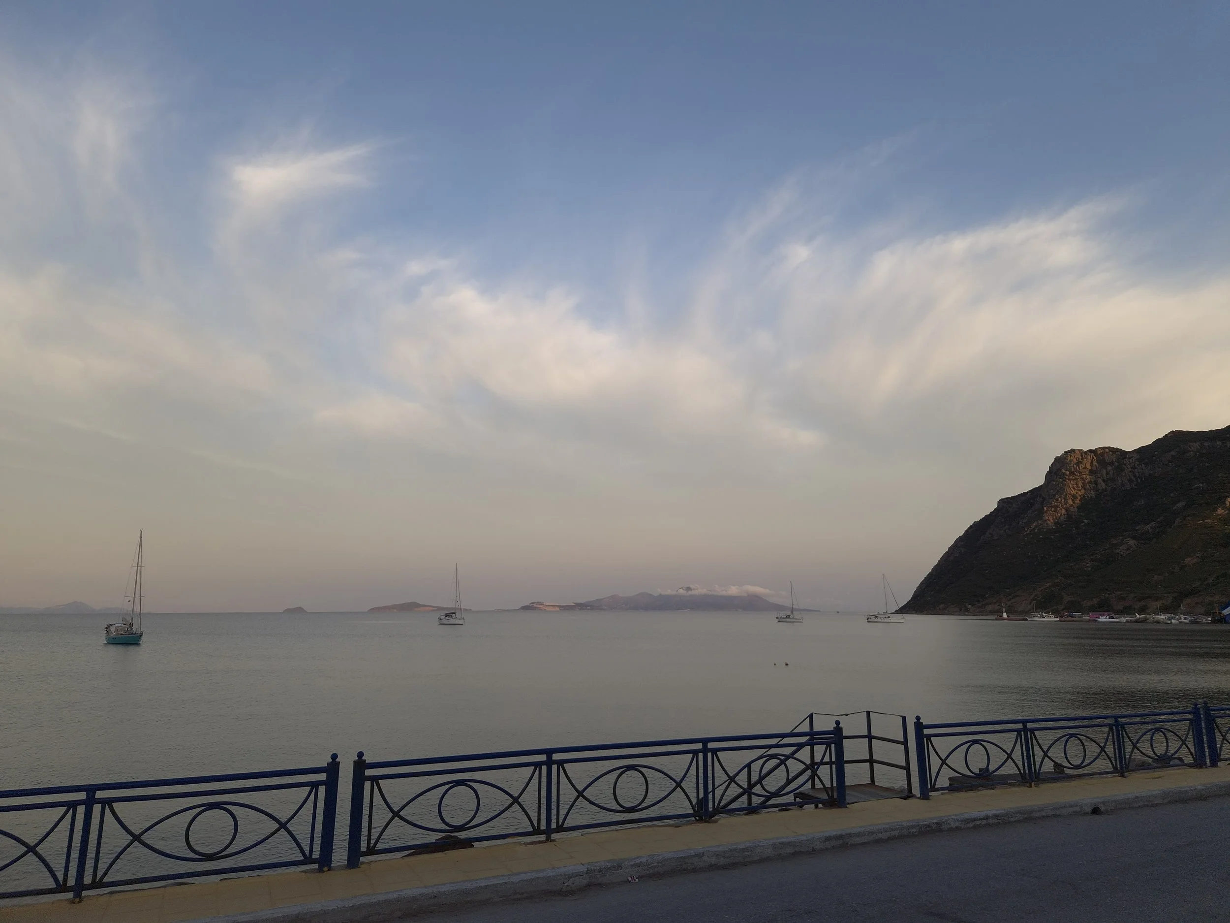 A tranquil waterfront scene with sailboats anchored in calm water, a mountainous landscape in the distance, and a sky with scattered clouds during the daytime. A decorative railing and a walkway are in the foreground.