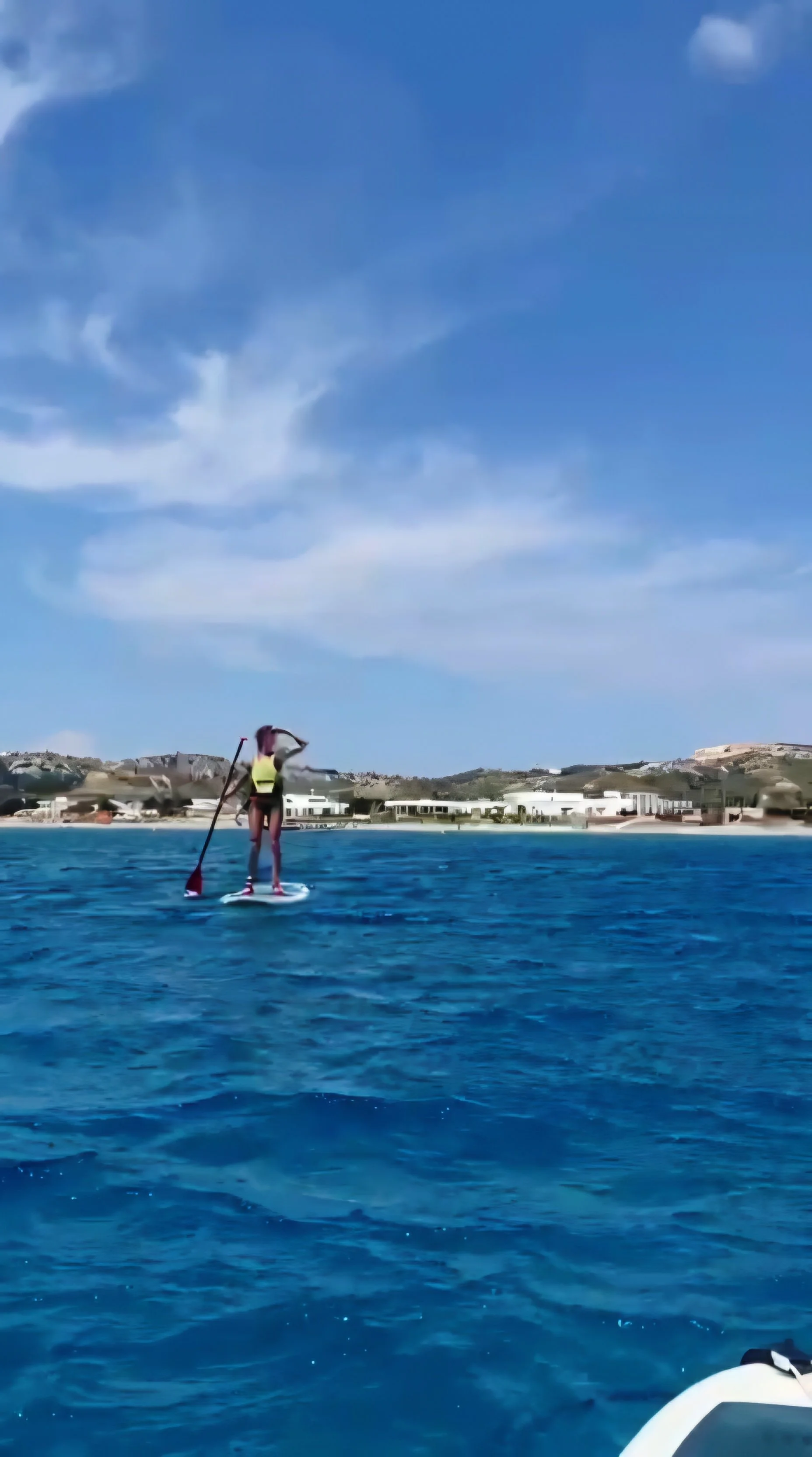 Person paddleboarding on the ocean with a bright blue sky and some clouds overhead, with buildings and hills in the background.