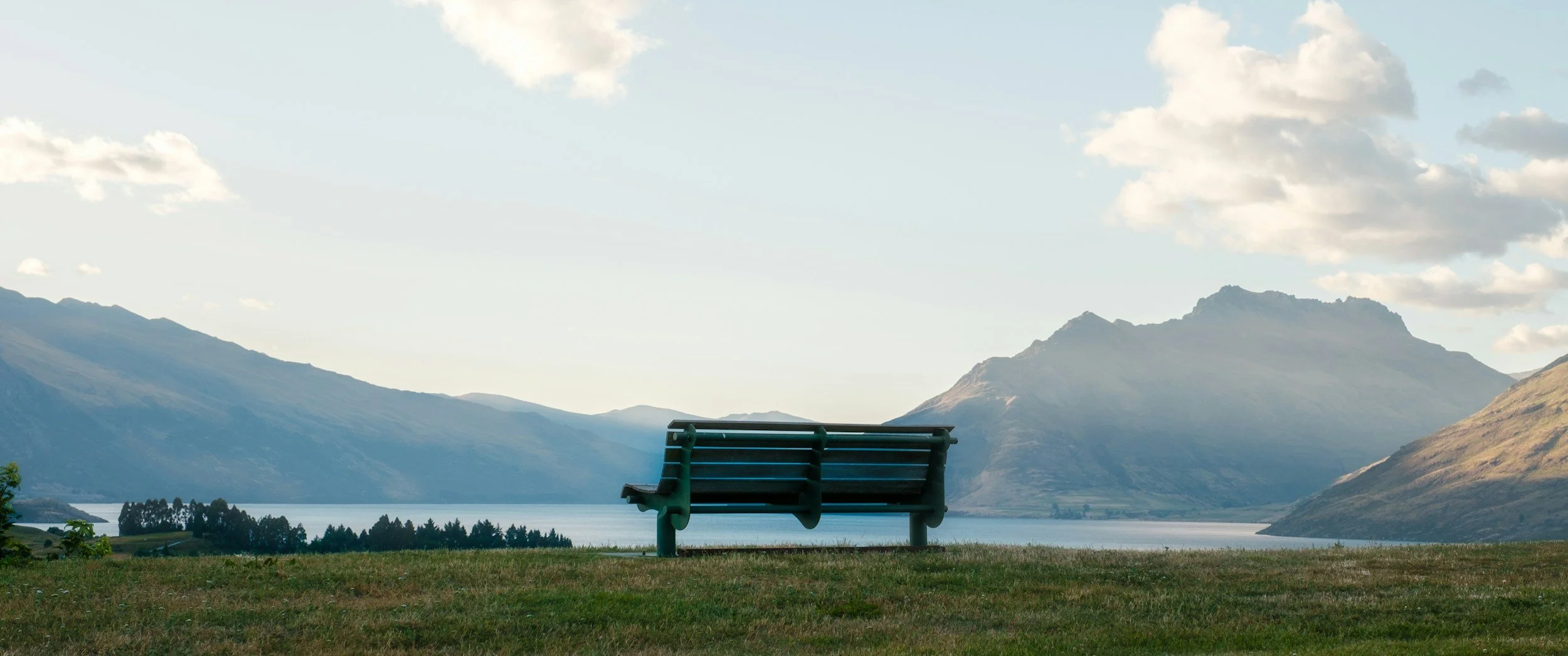 Empty bench overlooking a lake surrounded by mountains under a partly cloudy sky.