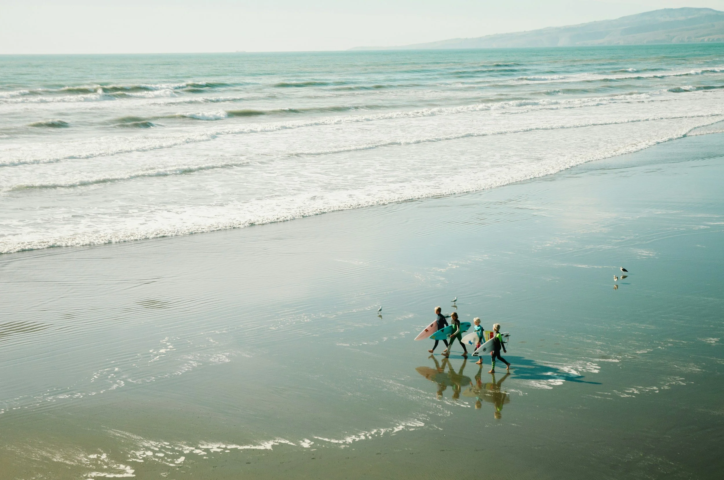 A group of children carrying surfboards walking along the beach near the water's edge, with seagulls nearby and waves approaching the shore.