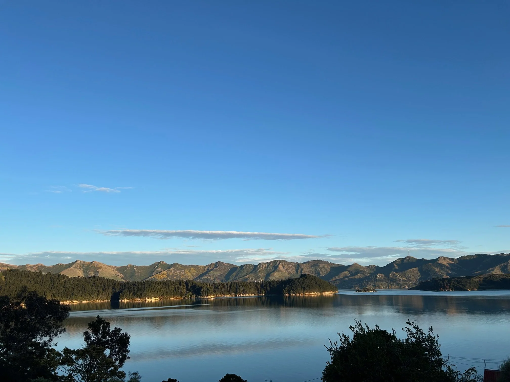 A scenic landscape featuring a serene lake with a calm water surface, bordered by lush green trees and mountains under a clear blue sky with a few scattered clouds.