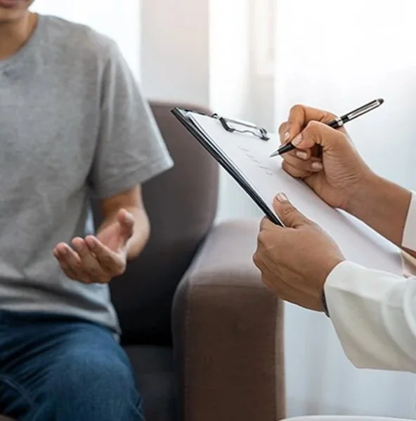 A person in a gray T-shirt and jeans speaking to a healthcare professional taking notes on a clipboard.
