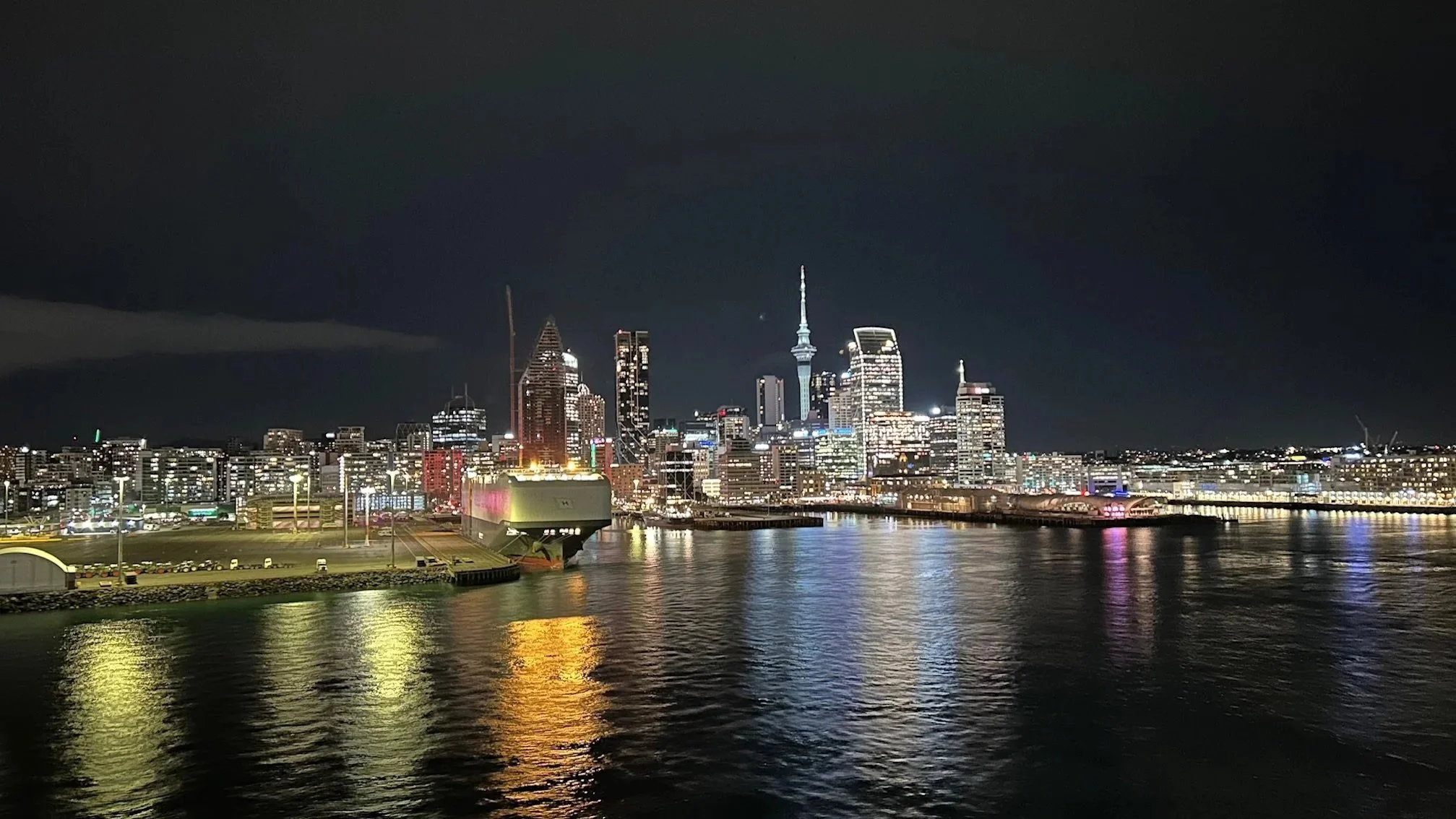 Nighttime view of a city skyline reflecting on a river, with a prominent tower, modern buildings, and illuminated skyscrapers.