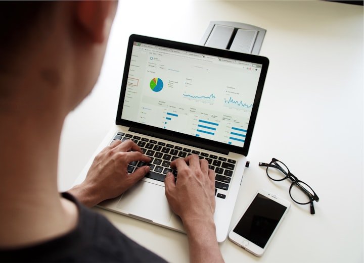 Person working on a laptop displaying data analytics, with glasses and a smartphone on a white desk.