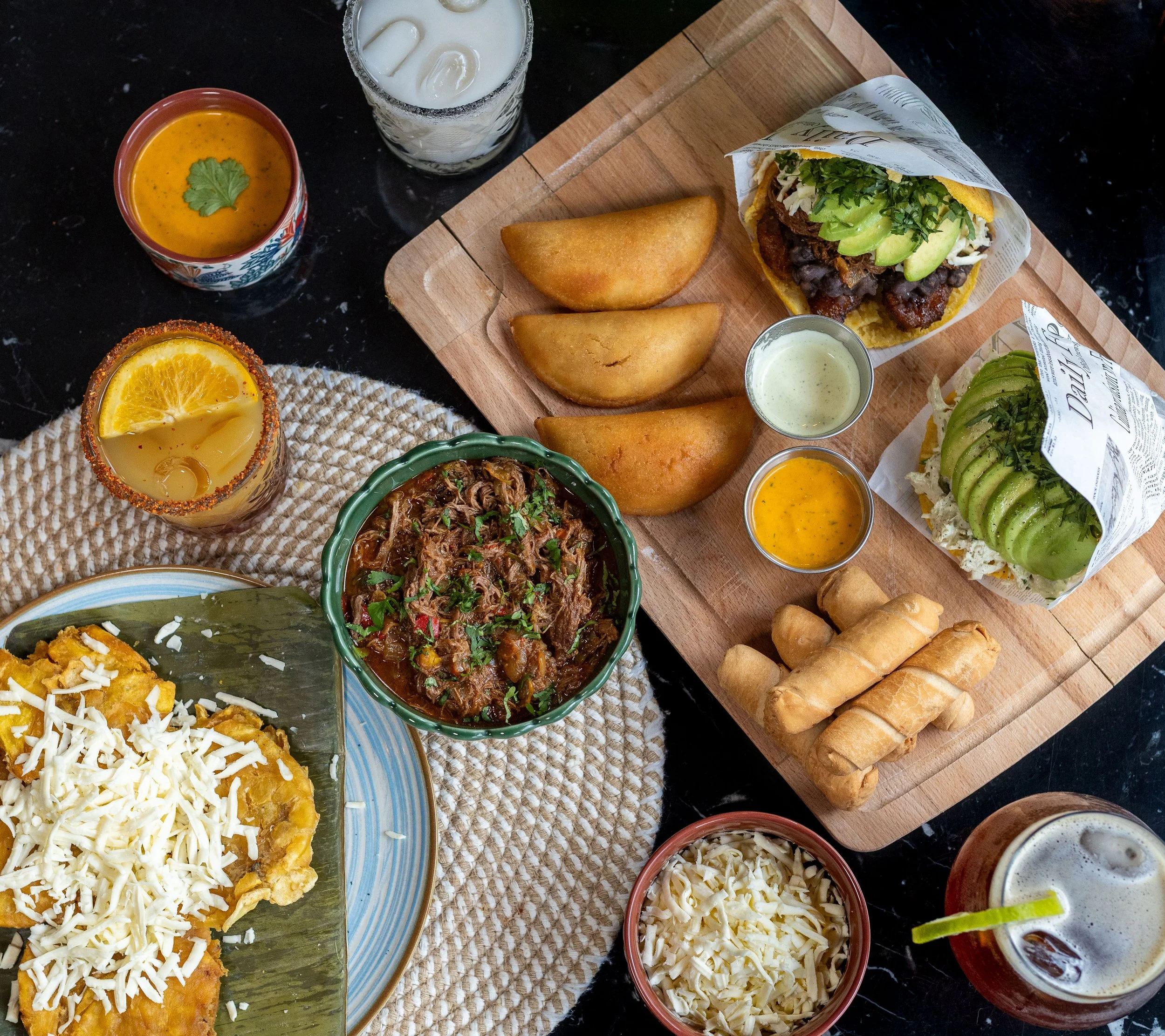 Mesa de comida venezolana con empanadas, tostones con queso blanco, tequeños, arepas y bebidas, sobre una mesa de mármol y un mantel de cuerdas.