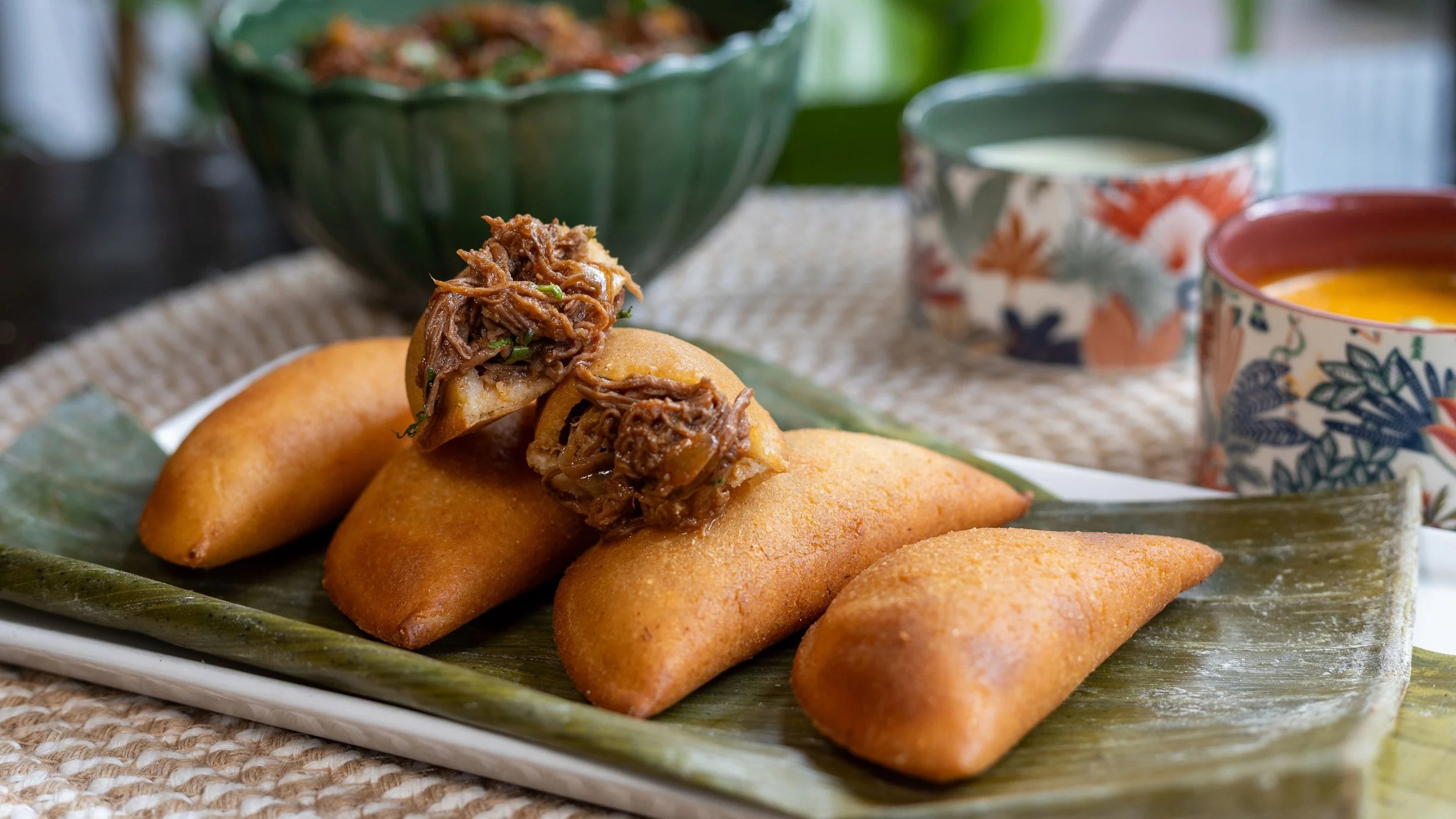 Empanadas de carne mechada en un plato con hojas de plátano, acompañadas de tazas con diferentes salsas.