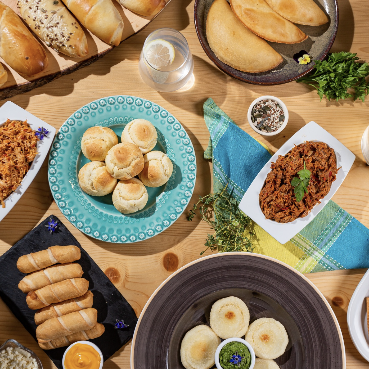 Diversos alimentos latinos tradicionales, incluyendo panes de bono, tequeños, carne mechada, empanadas, cachitos, salsas, y acompañamientos, sobre una mesa de madera decorada con manteles y flores comestibles.