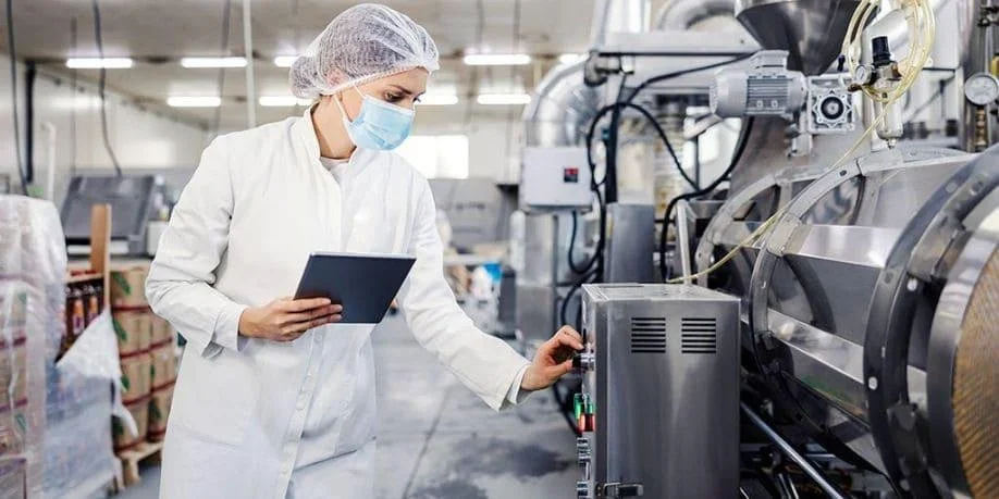 Mujer en un laboratorio de producción alimentaria operando maquinaria y revisando un dispositivo, con un uniforme de protección, máscara facial y gorro.