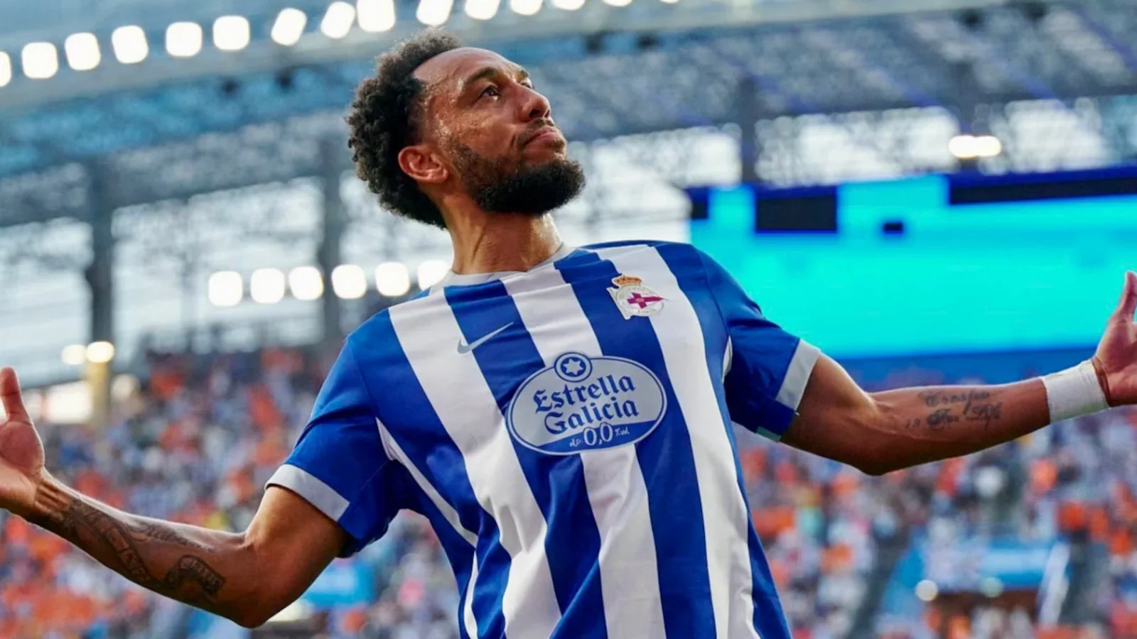 A soccer player wearing a blue and white striped jersey with a crest and sponsor logo, celebrating on the field with arms outstretched and a look of triumph.