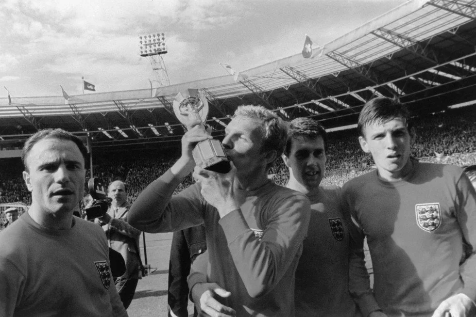 Four soccer players wearing England national team jerseys celebrating on a soccer field; one is kissing a trophy.
