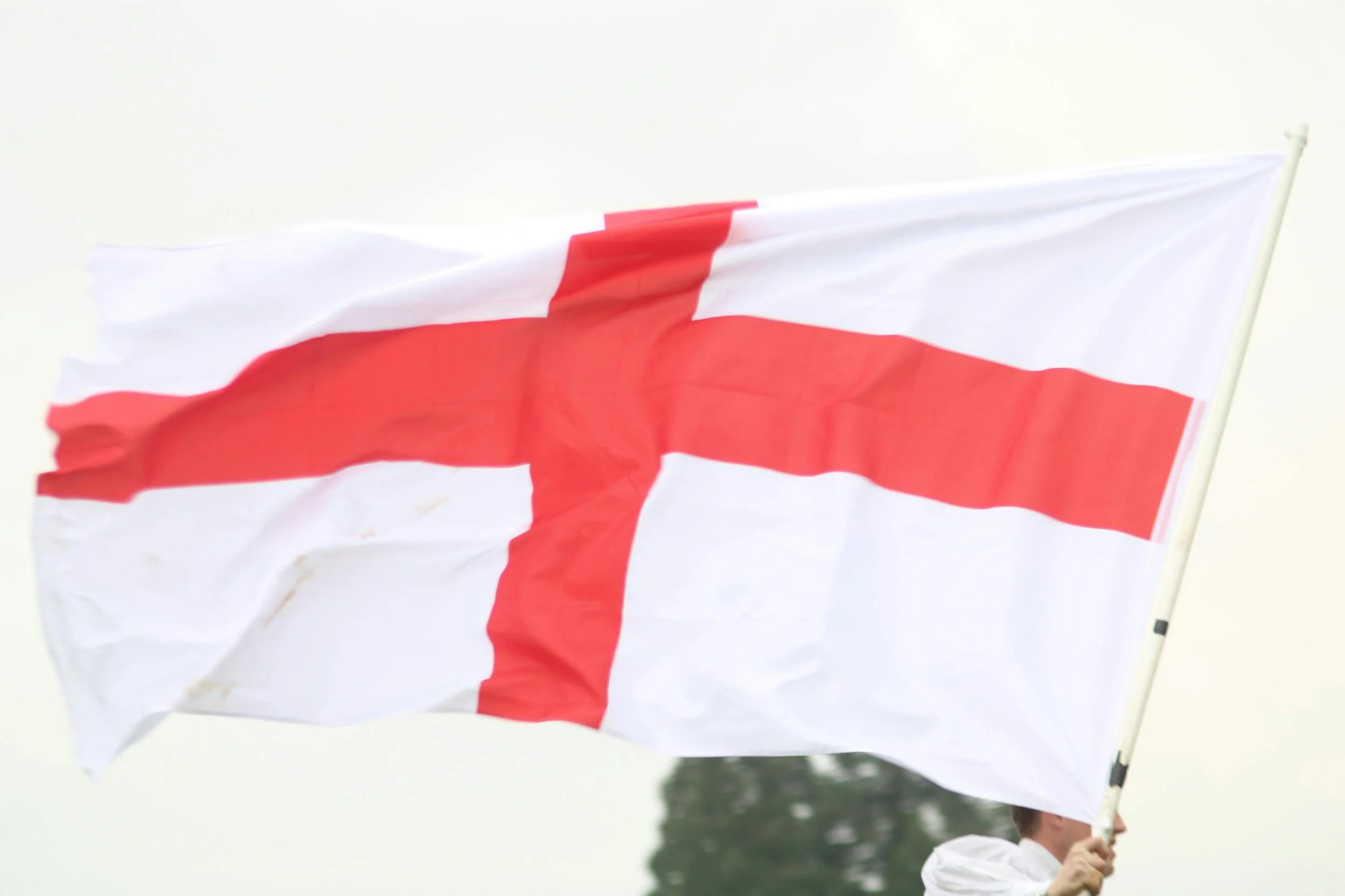 A person holding an England flag with a red cross on a white background, outdoors on a cloudy day.