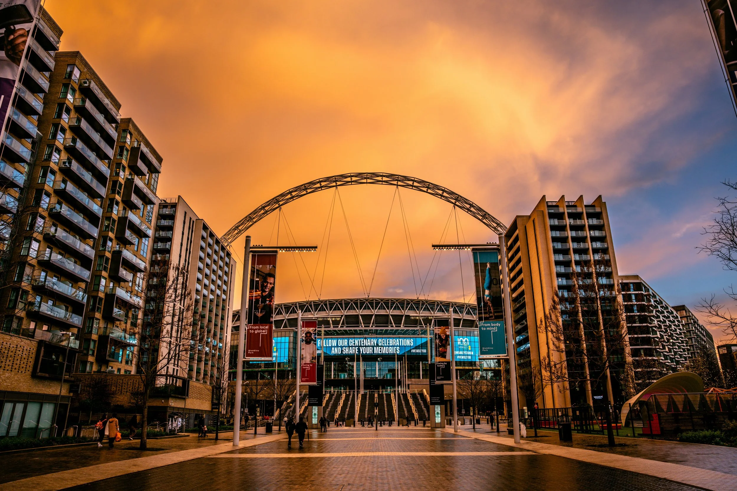 Wide shot of a city square at sunset with modern multi-story apartment buildings on either side and a large stadium or arena in the center. The sky is orange with clouds and there are a few pedestrians walking.