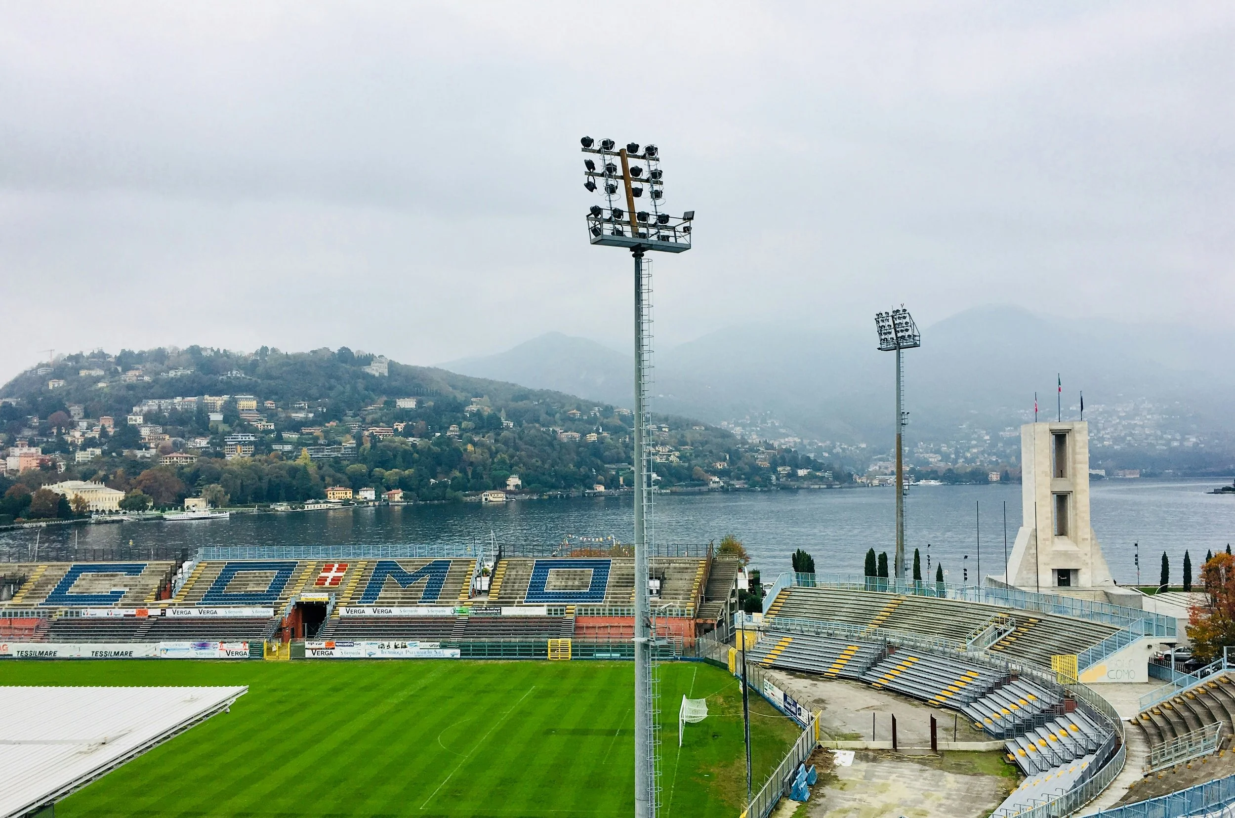 Empty stadium with a green field and colorful seats, surrounded by a track, near a river with a hillside town in the background and floodlights above the field.
