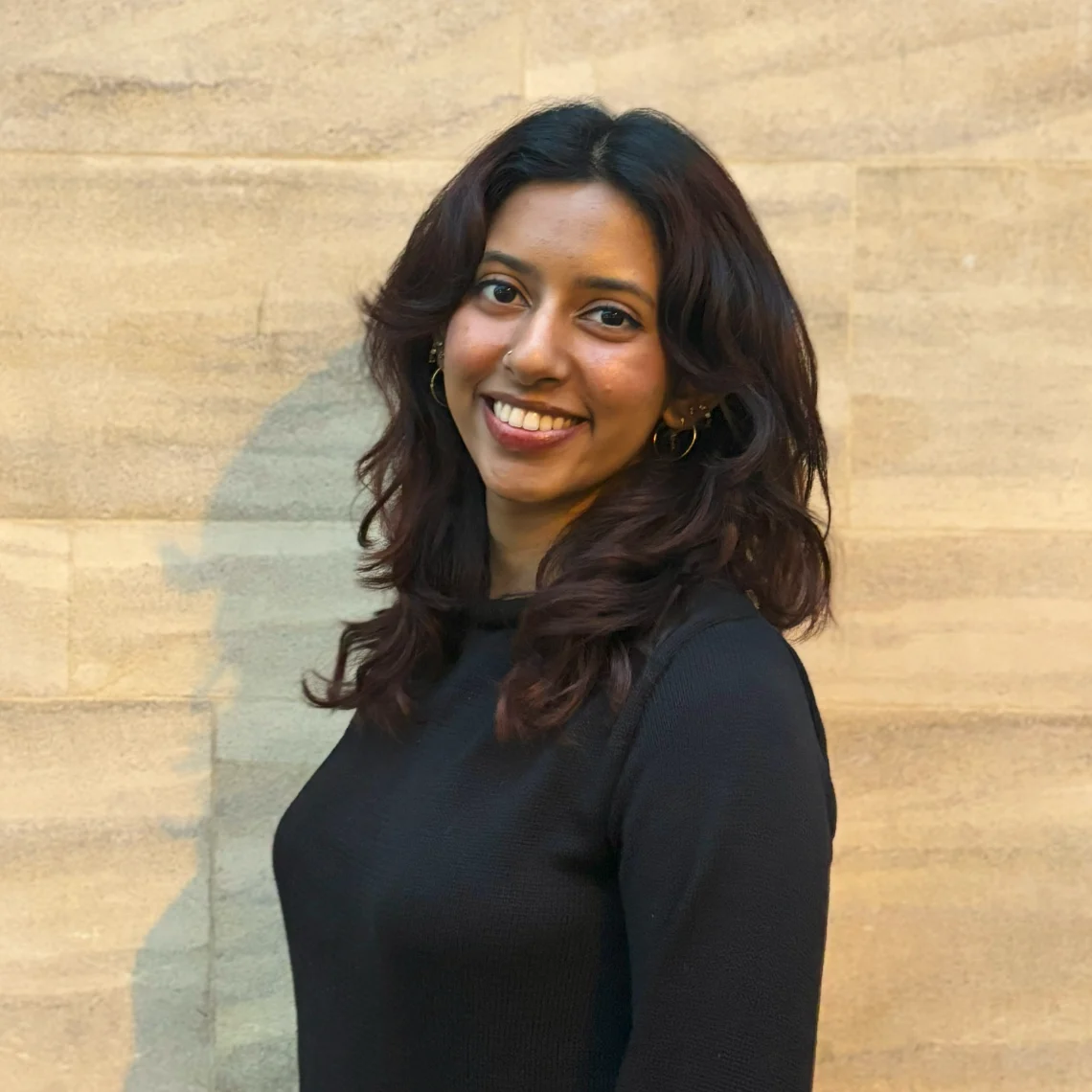A smiling woman with wavy dark hair, earrings, and a nose ring, wearing a black top, standing against a light brown textured wall.