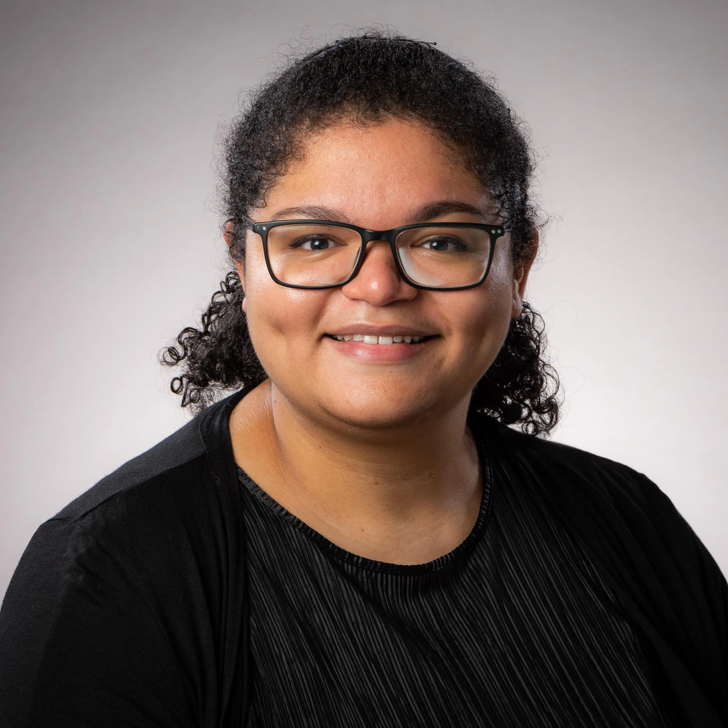A woman with curly hair wearing glasses, smiling, dressed in a black top with pleated fabric detail.