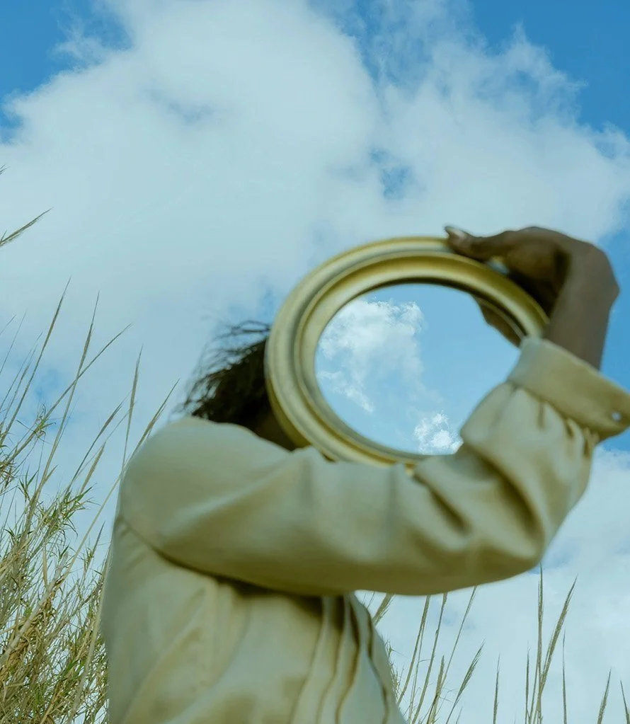 Person holds a round mirror up to the sky, reflecting clouds, with tall grass and a partly cloudy sky background.