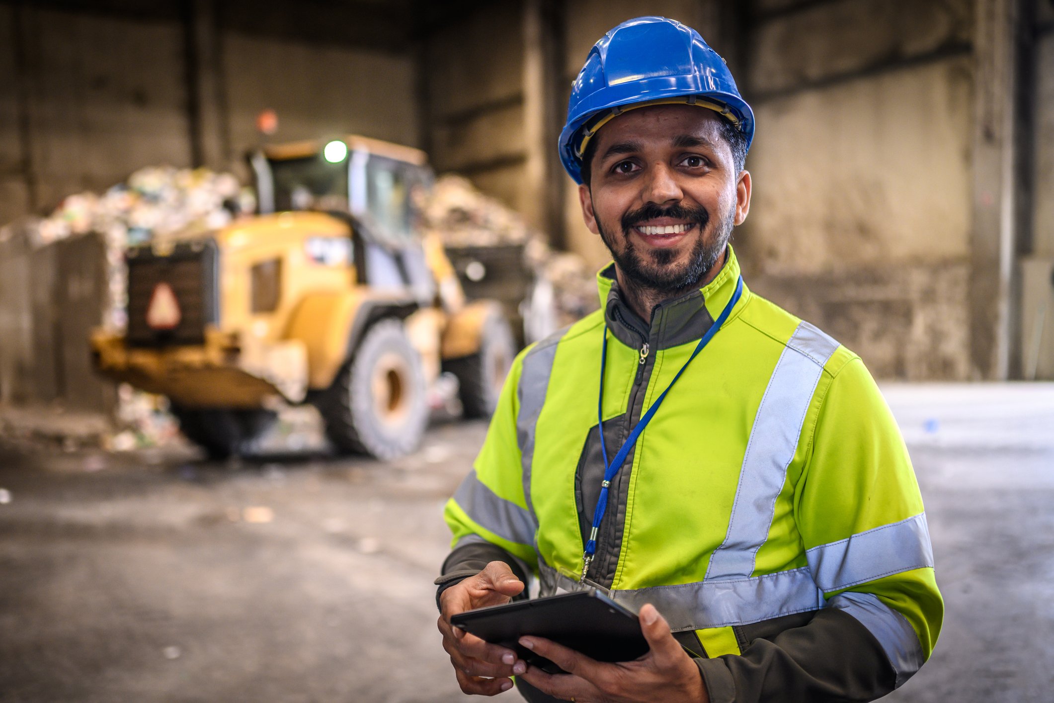 Happy man at waste facility with an Ipad, wearing high vis and helmet