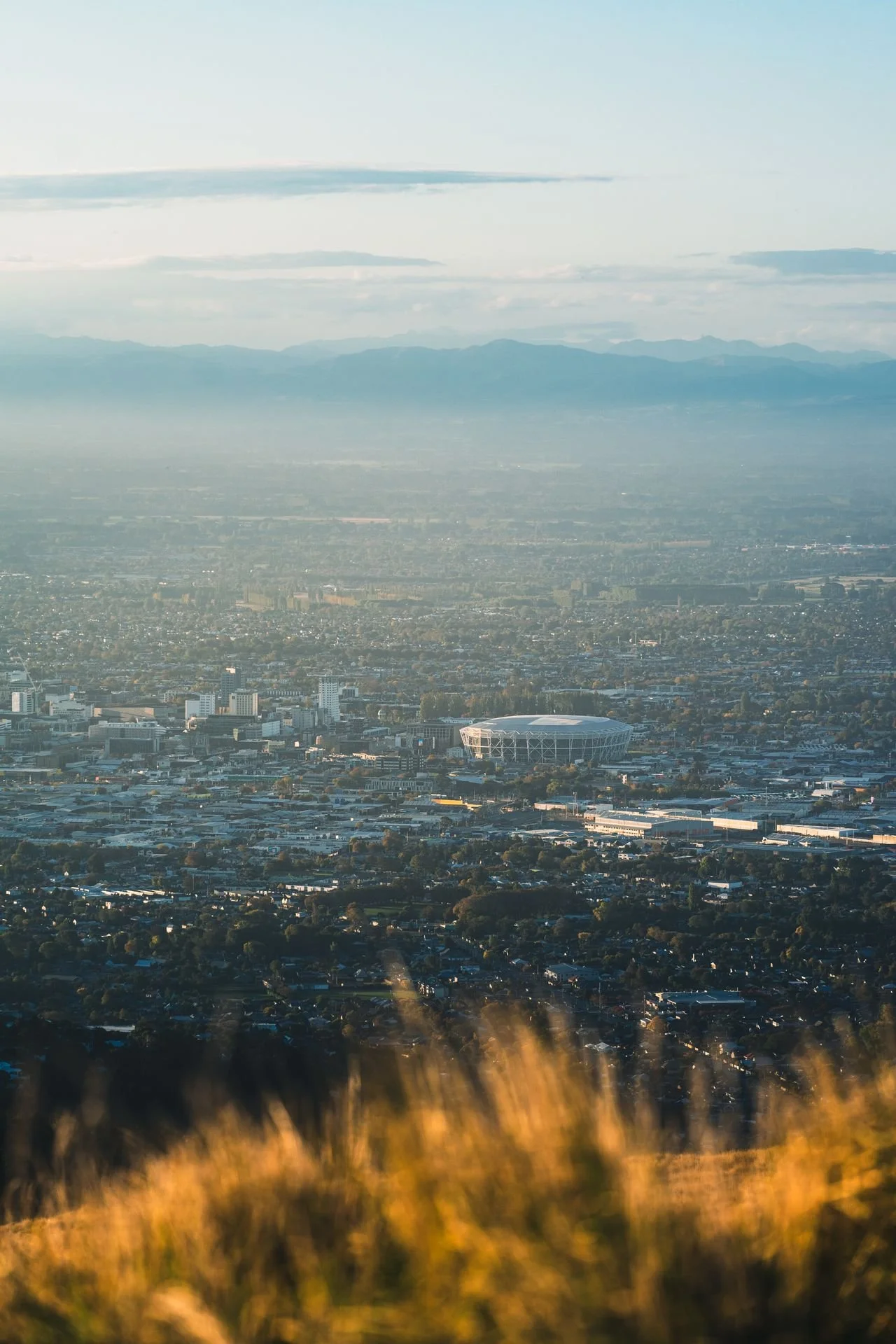 Aerial view of a cityscape with a stadium, mountains in the background, and a hazy sky.