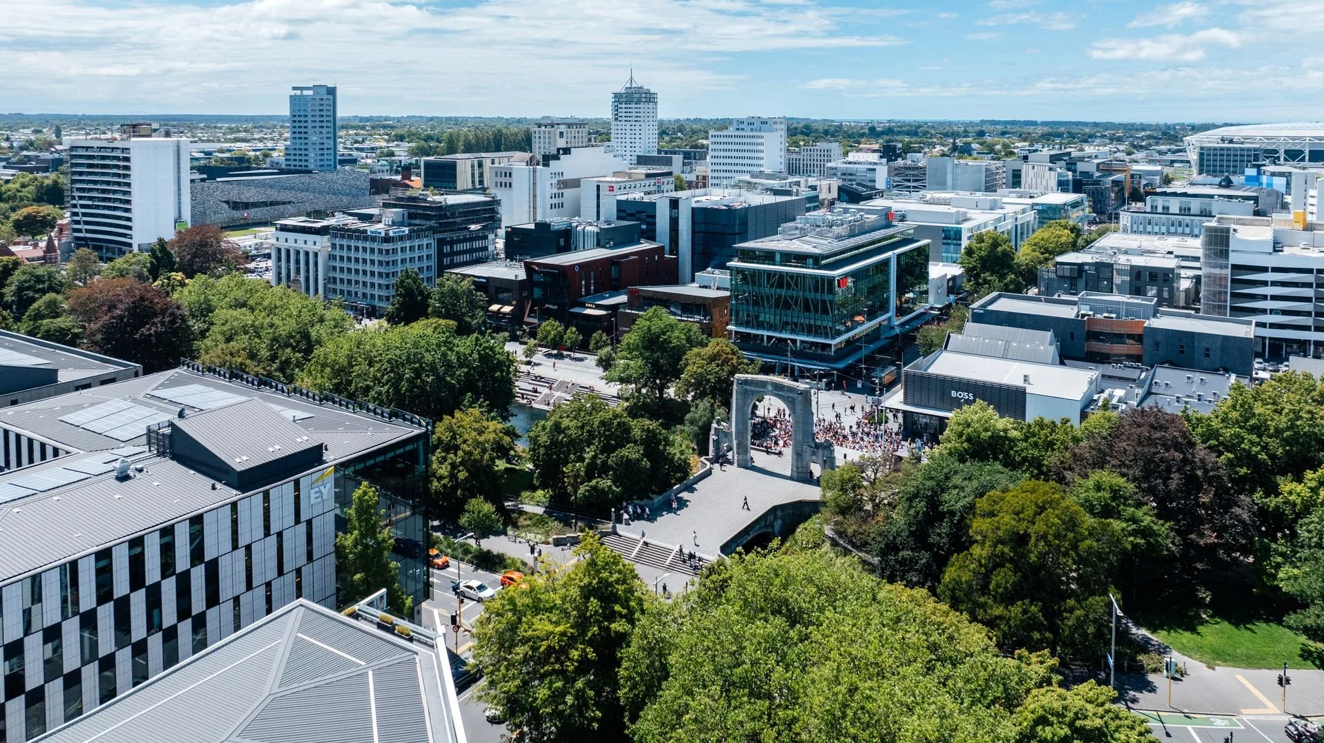 Aerial view of a Christchurch cityscape with modern buildings, green trees, and a crowd gathered near a historical arch monument.