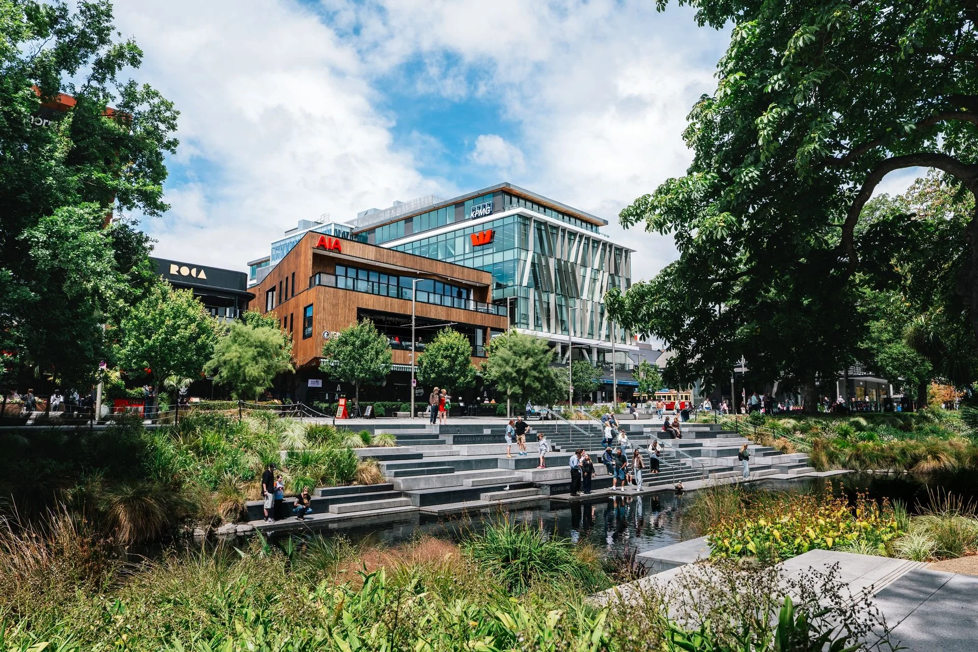 People sitting and walking on steps beside a water feature in a city park, with modern office buildings and trees in the background.