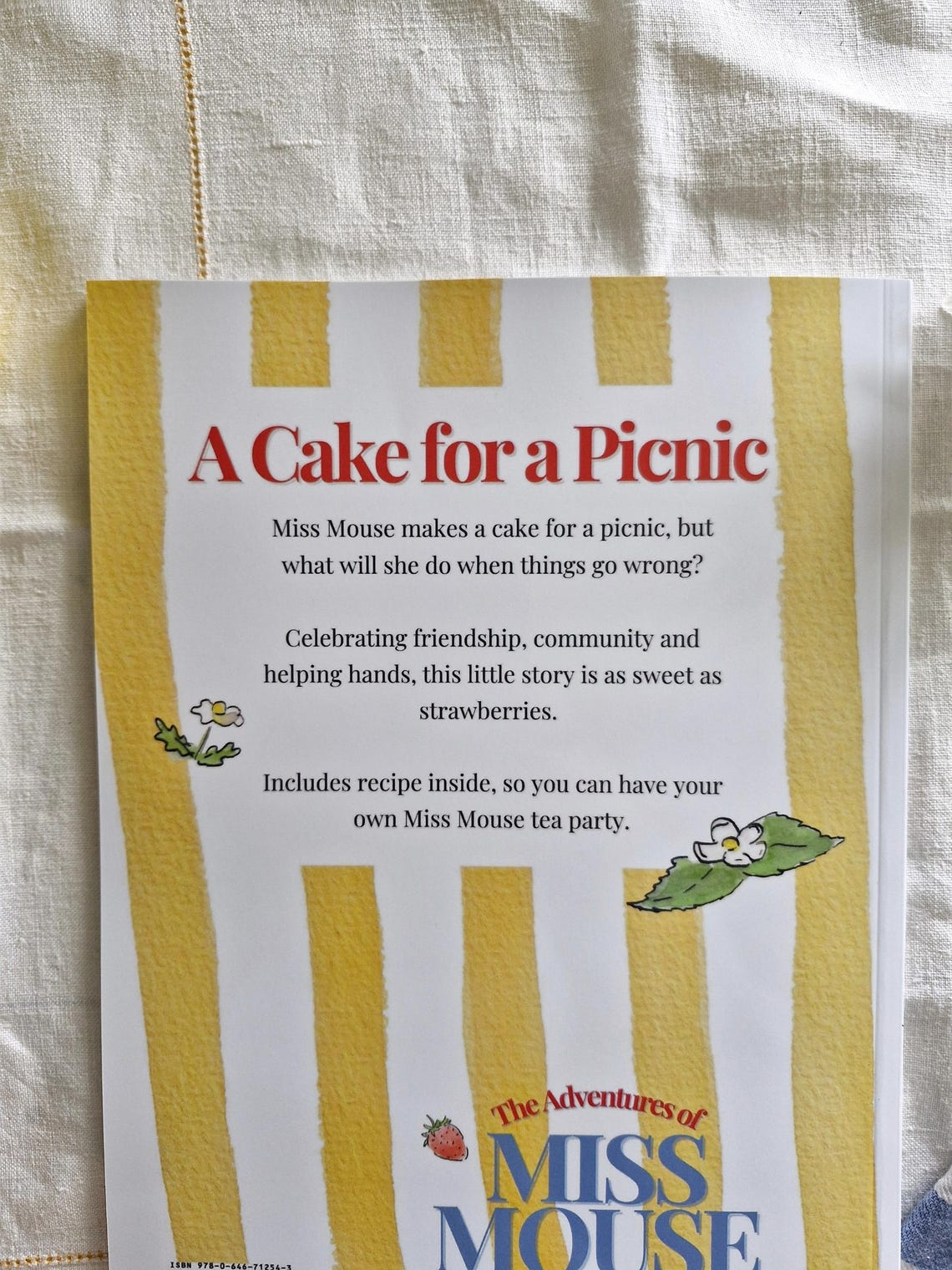 A book cover titled "A Cake for a Picnic" featuring illustrations of strawberries and floral decorations, with a beige fabric background.