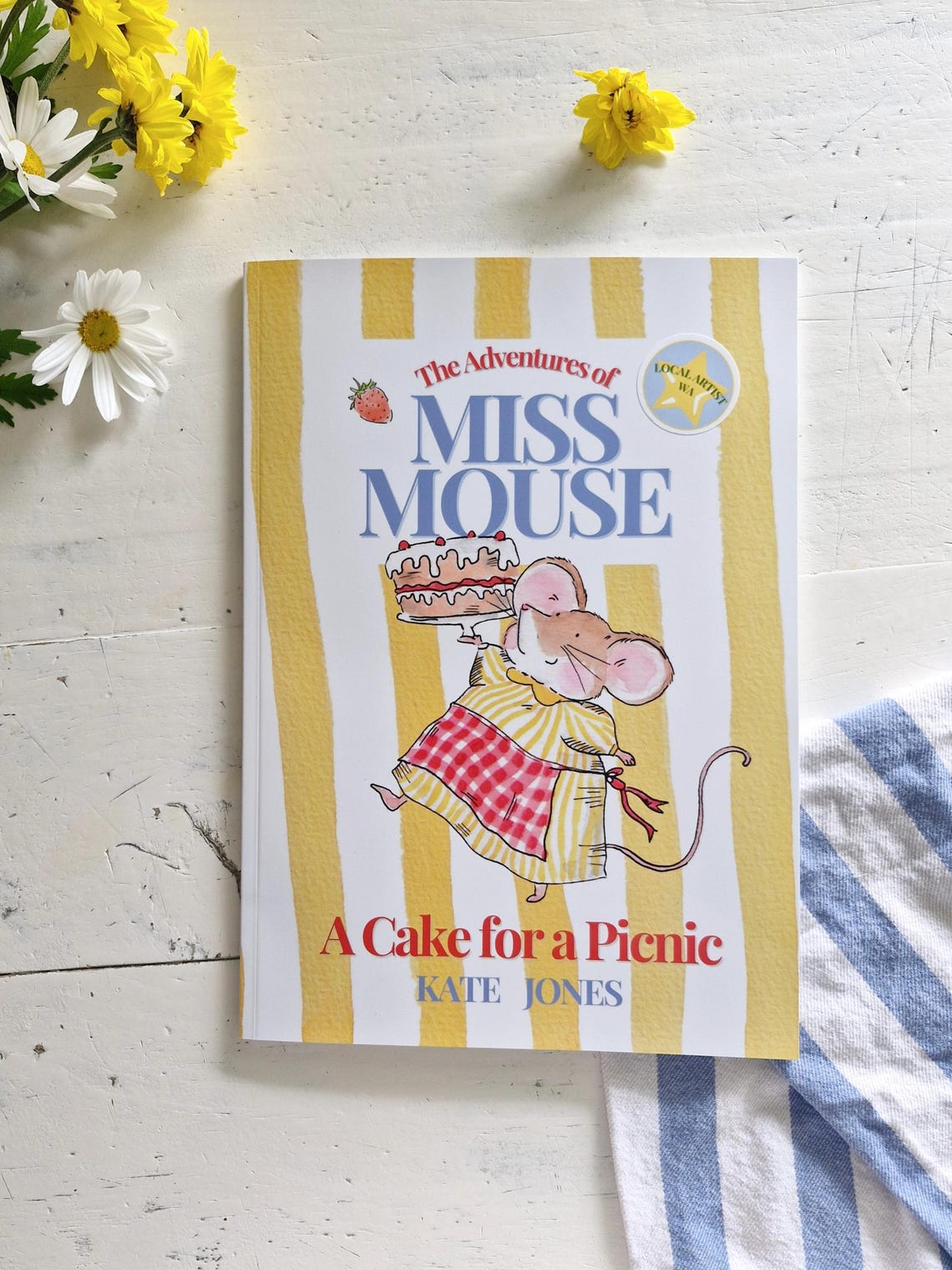 Book titled "The Adventures of Miss Mouse: A Cake for a Picnic" by Kate Jones, with an illustration of a mouse carrying a cake, placed on a white surface with yellow and white striped background, surrounded by yellow and white flowers and a blue stri